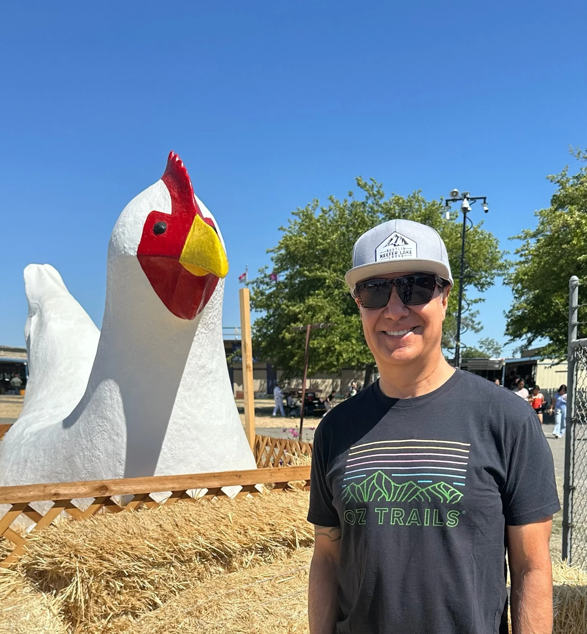 Man smiling in front of a large chicken sculpture outdoors