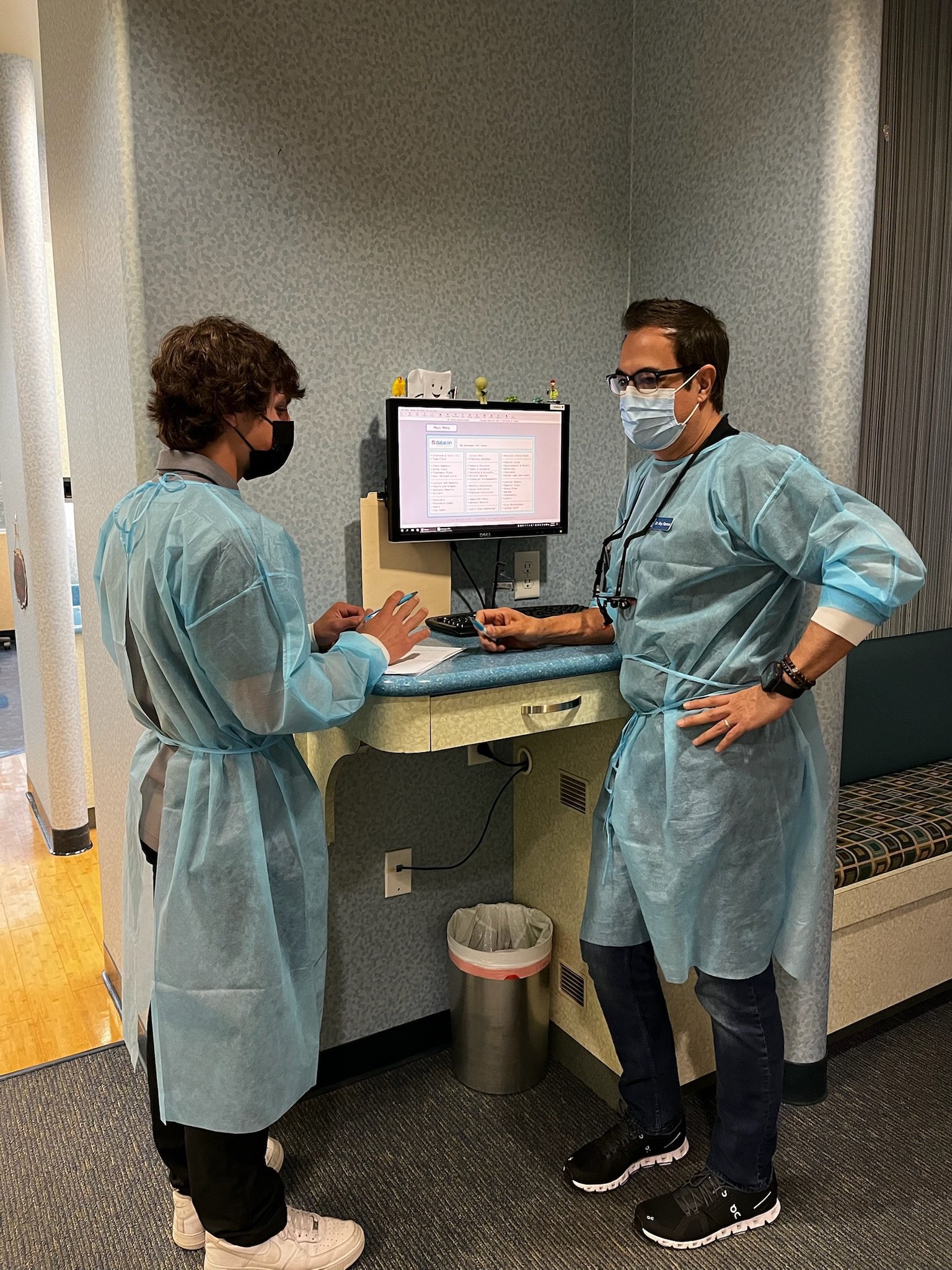 Two people in masks and disposable gowns talking beside a computer monitor in a dental office