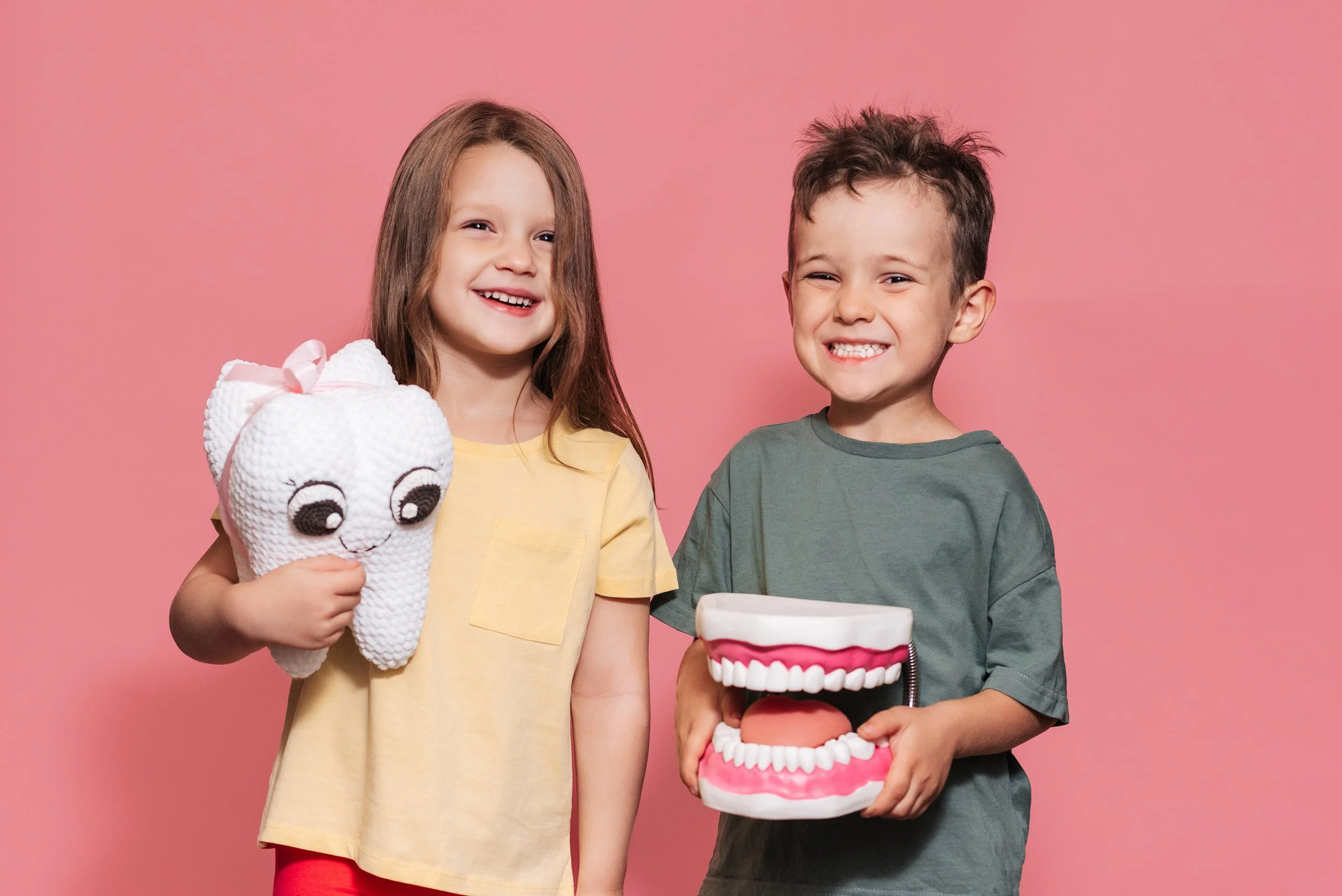 Two children smiling, holding a tooth plush and a dental model