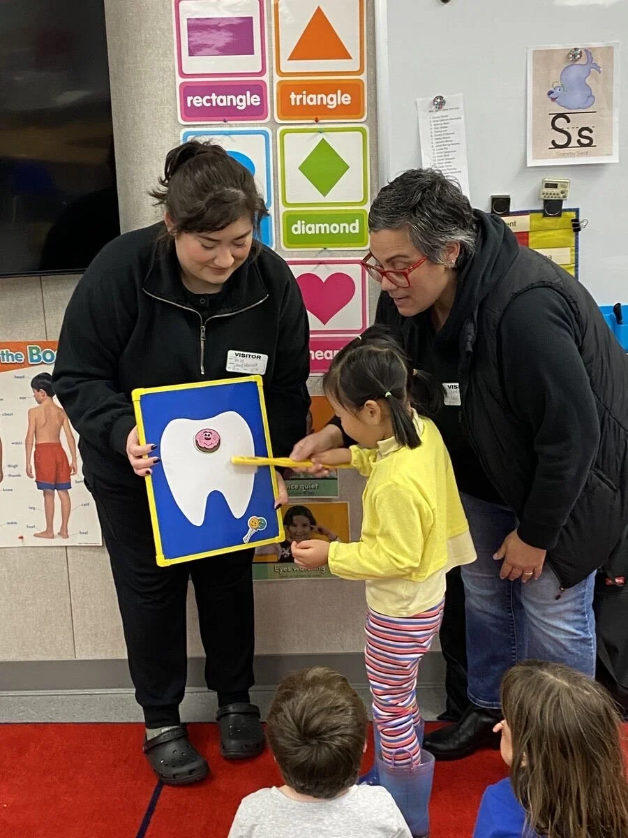 Child pointing to a tooth diagram while two adults assist in a classroom