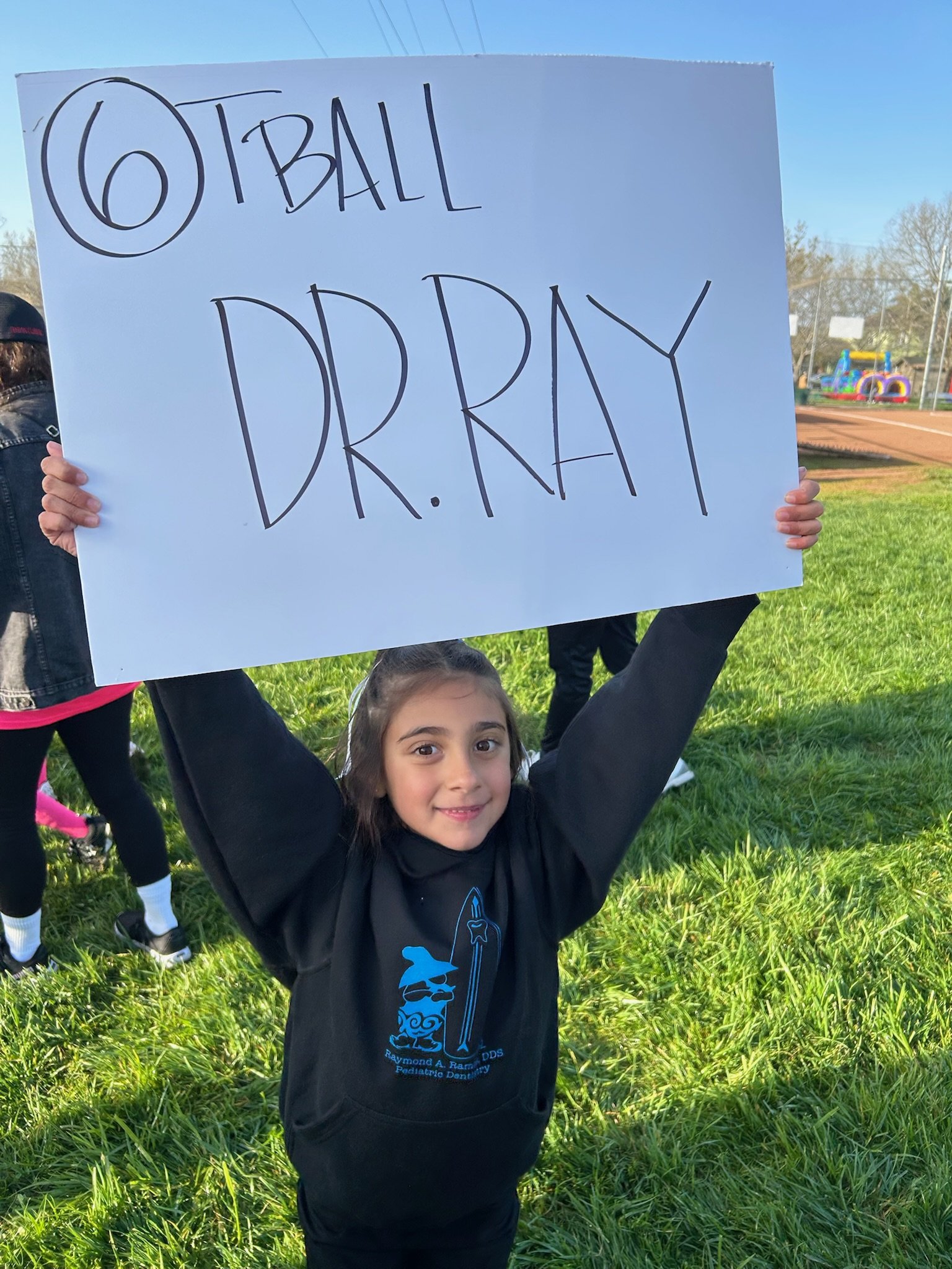 Child holding a large sign that reads ‘OTBALL DR. RAY’ on a grassy field