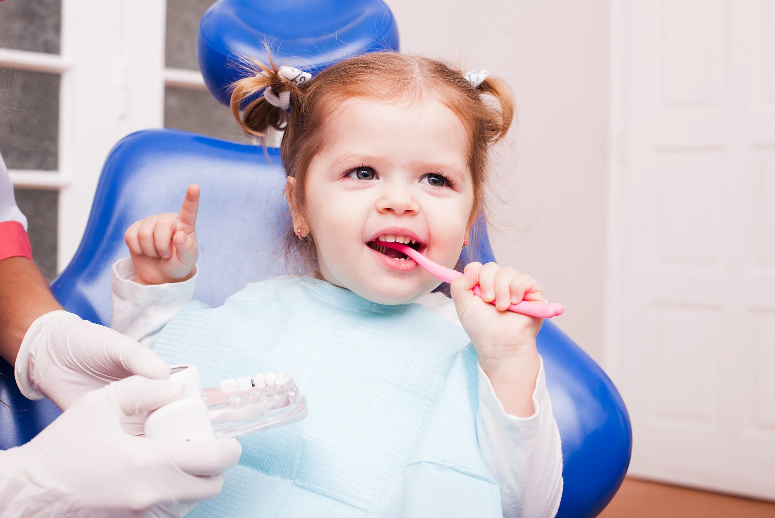 young toddler smiling in the dental chair brushing her teeth with a pink toothbrush and holding up her other hand happily
