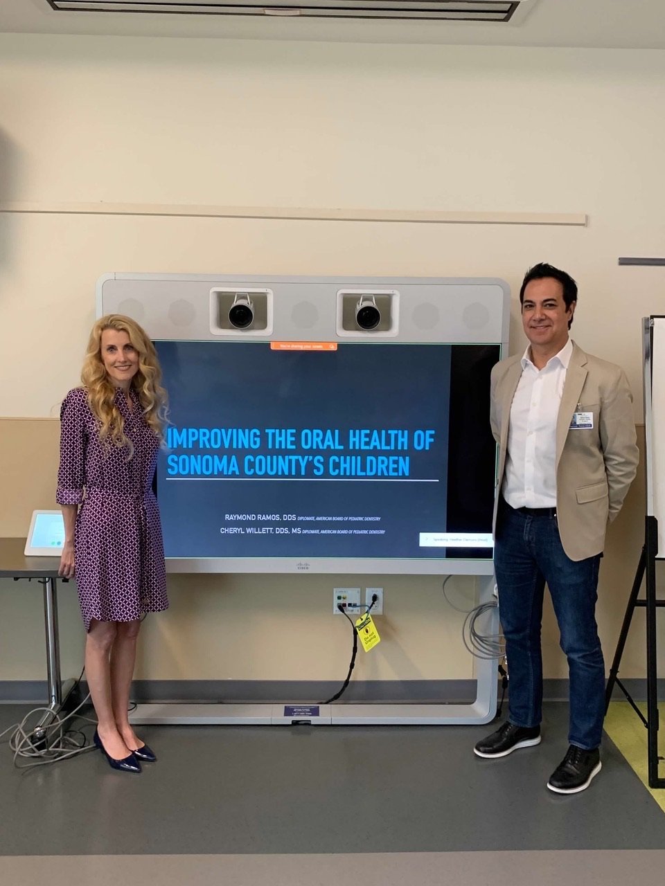 Two adults standing beside a large display screen that reads ‘Improving the Oral Health of Sonoma County’s Children