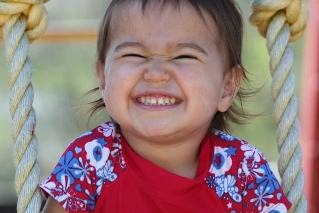 A young girl smiling happily on a swing, wearing a red shirt with blue and white floral patterns.