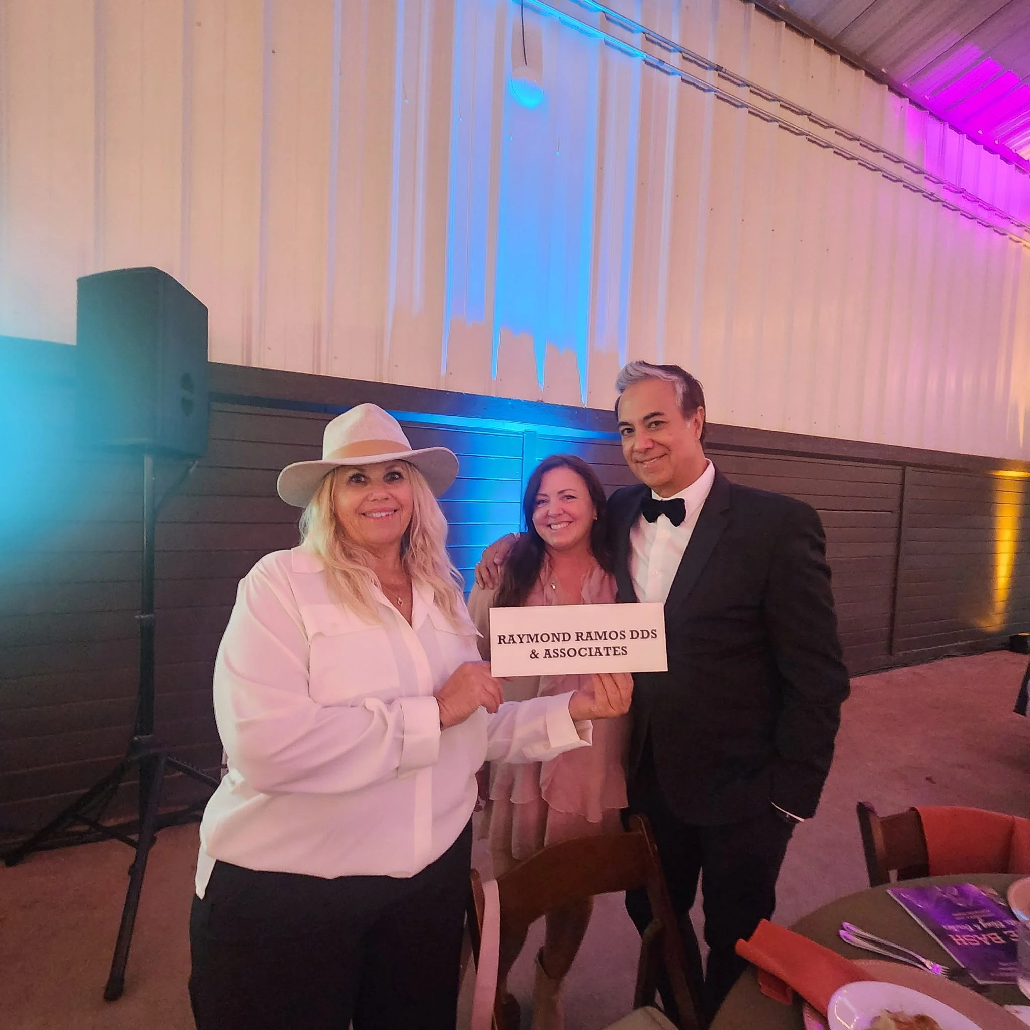 Three people posing indoors; one holds a sign that reads ‘RAYMOND RAMOS DDS & ASSOCIATES