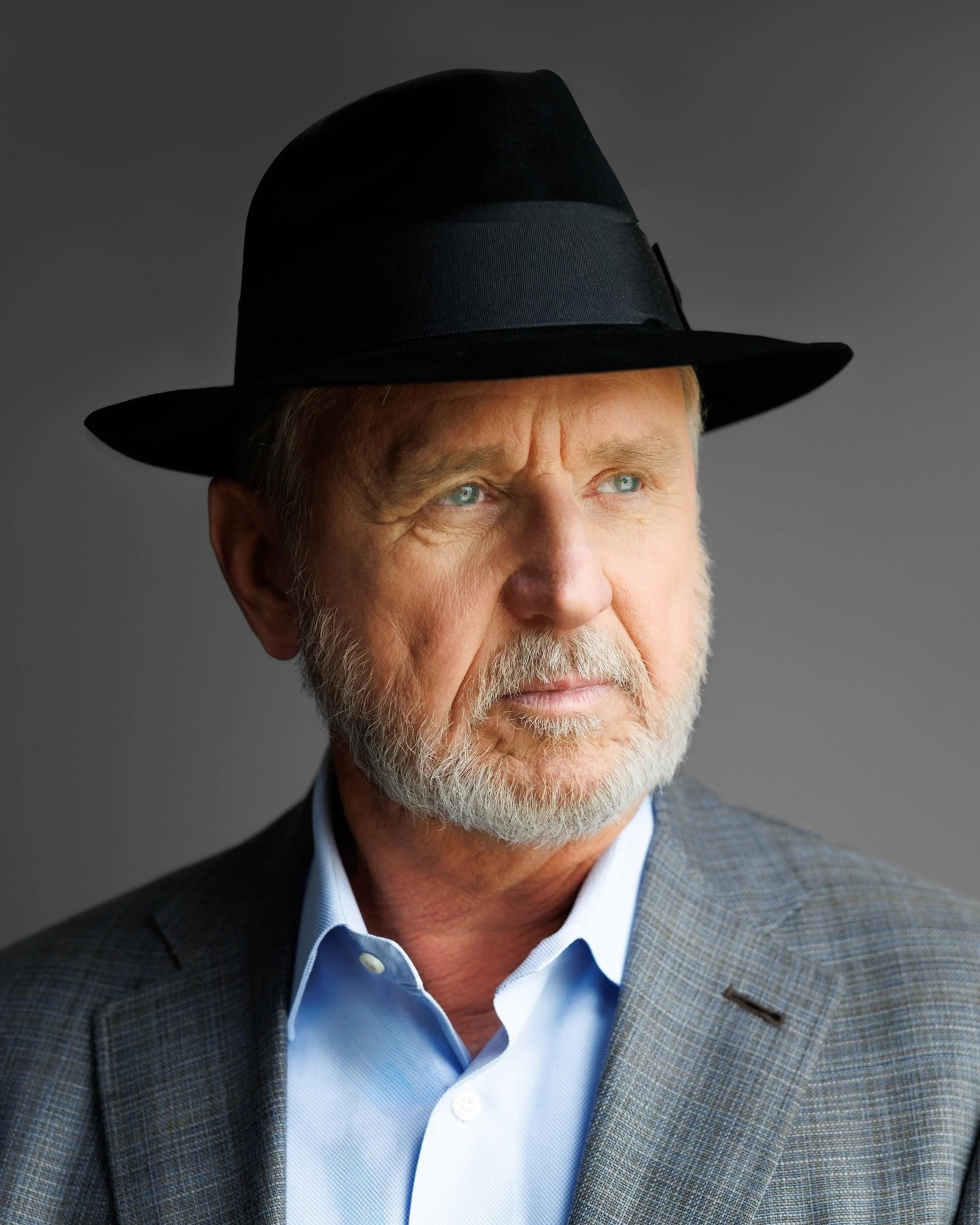 Ray Tye is a mature man with a gray beard and blue eyes wearing a gray blazer, a light blue dress shirt, and a black fedora hat, looking off to the side against a neutral background.