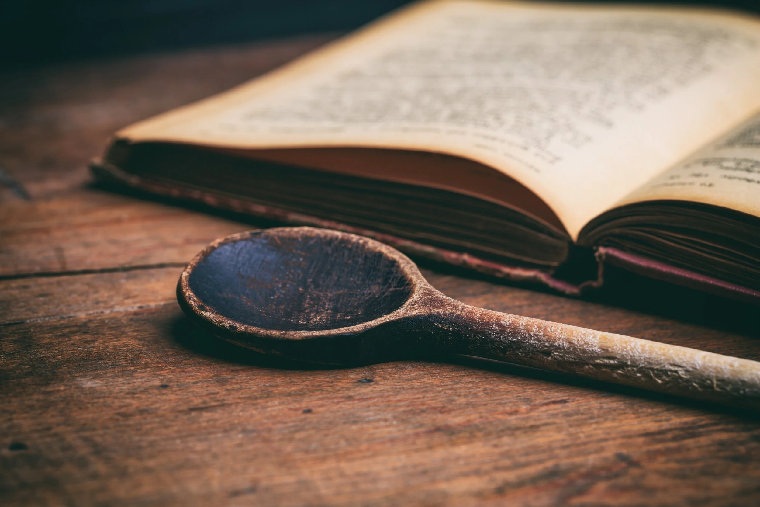 Close-up of a wooden spoon next to an open book on a wooden table.