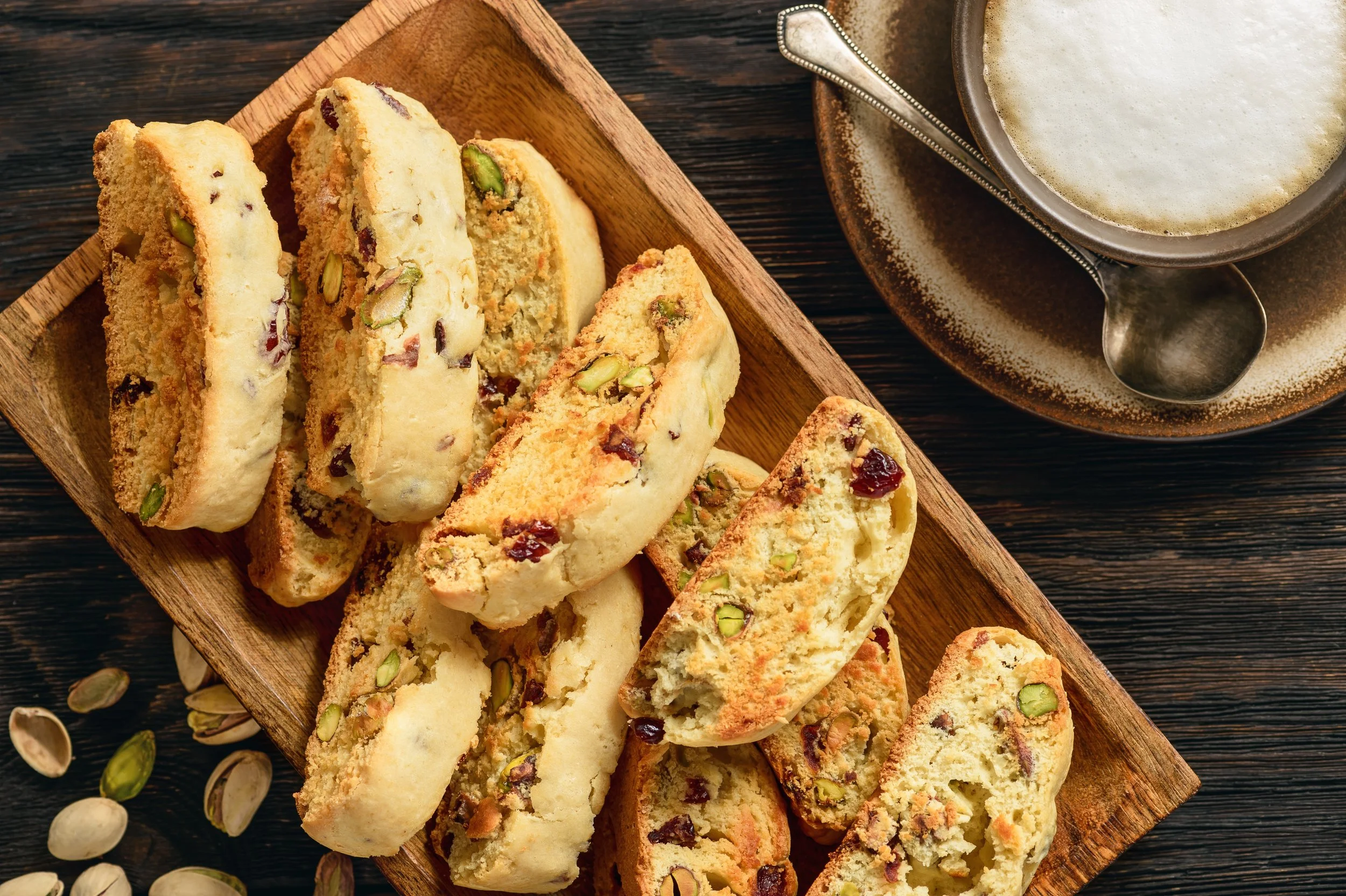 Biscotti with pistachios and cranberries in a wooden tray next to a cup of cappuccino on a rustic table.