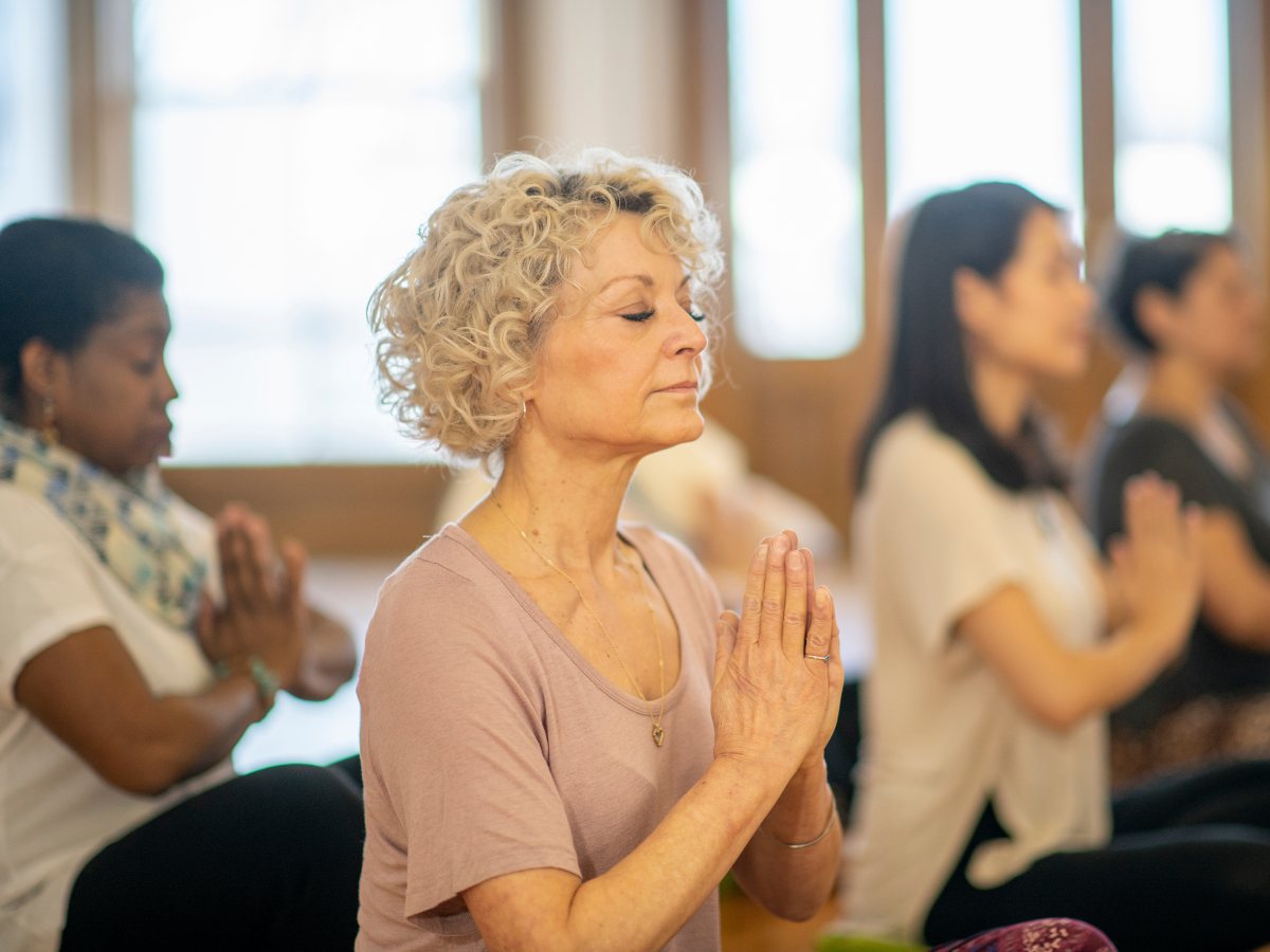 A yoga class is seated on their mats, relaxed, eyes closed, with their hands at their hearts.