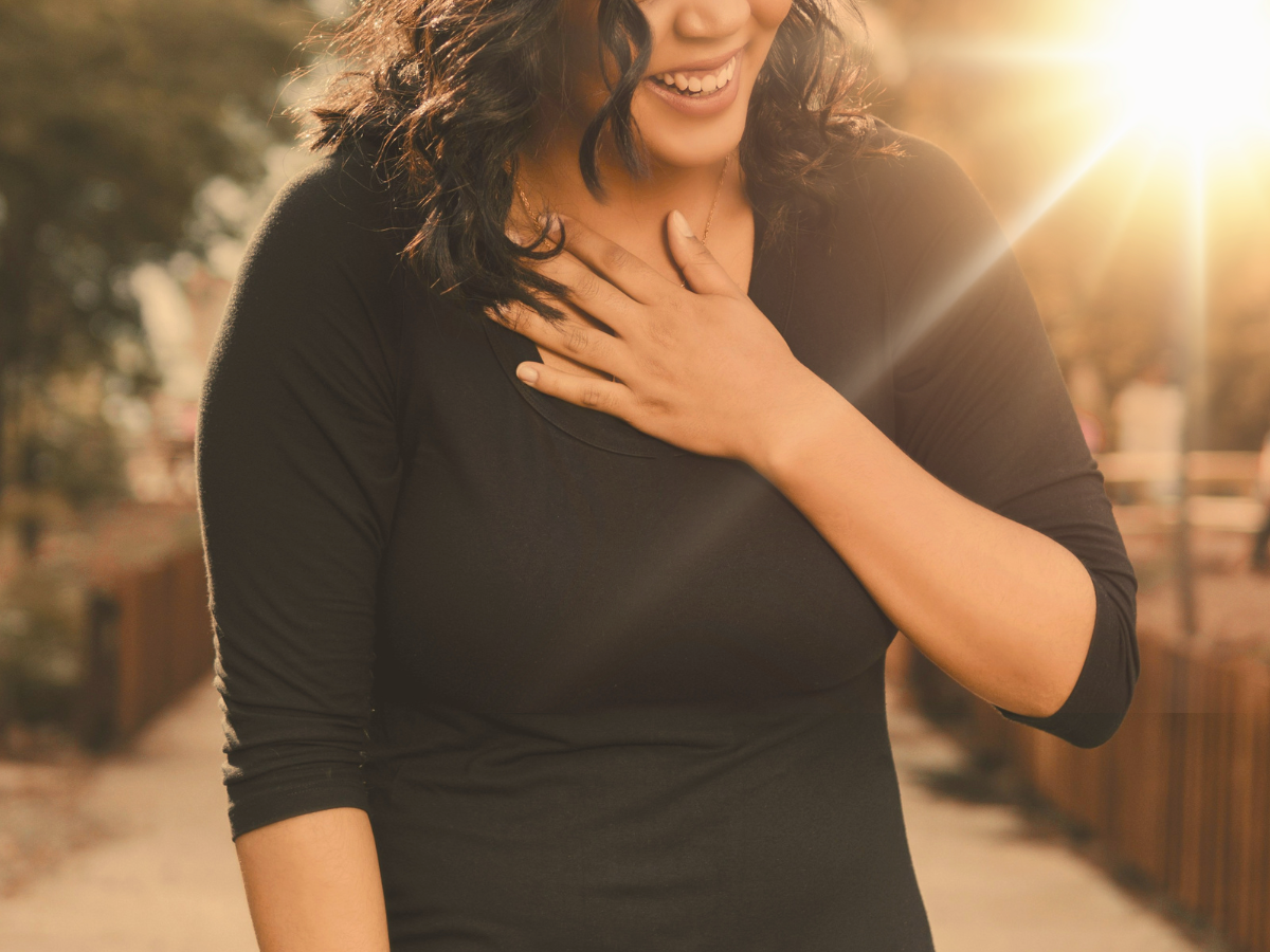 A woman stands in the rays of the sun with her hand on her heart.  She is smiling with joy.