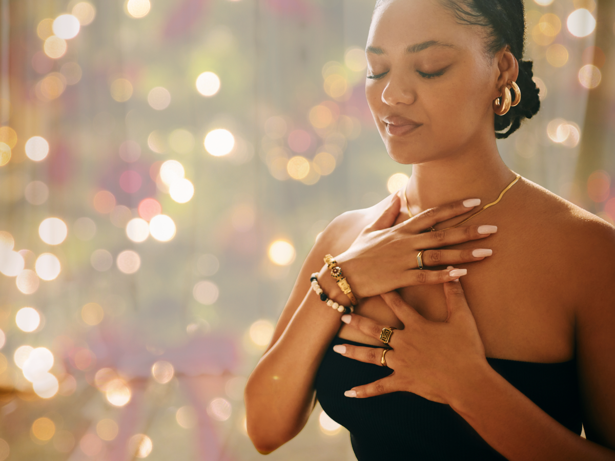 woman puts hand on heart with eyes closed in front of a background of holiday lights.