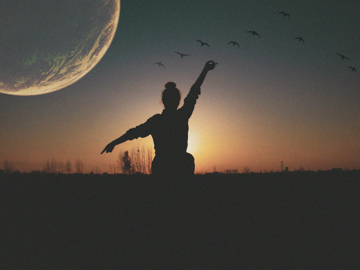 A woman stands in silhouette with arms extended in a field in front of a setting sun and rising moon.  A flock of birds flies free in the sky.