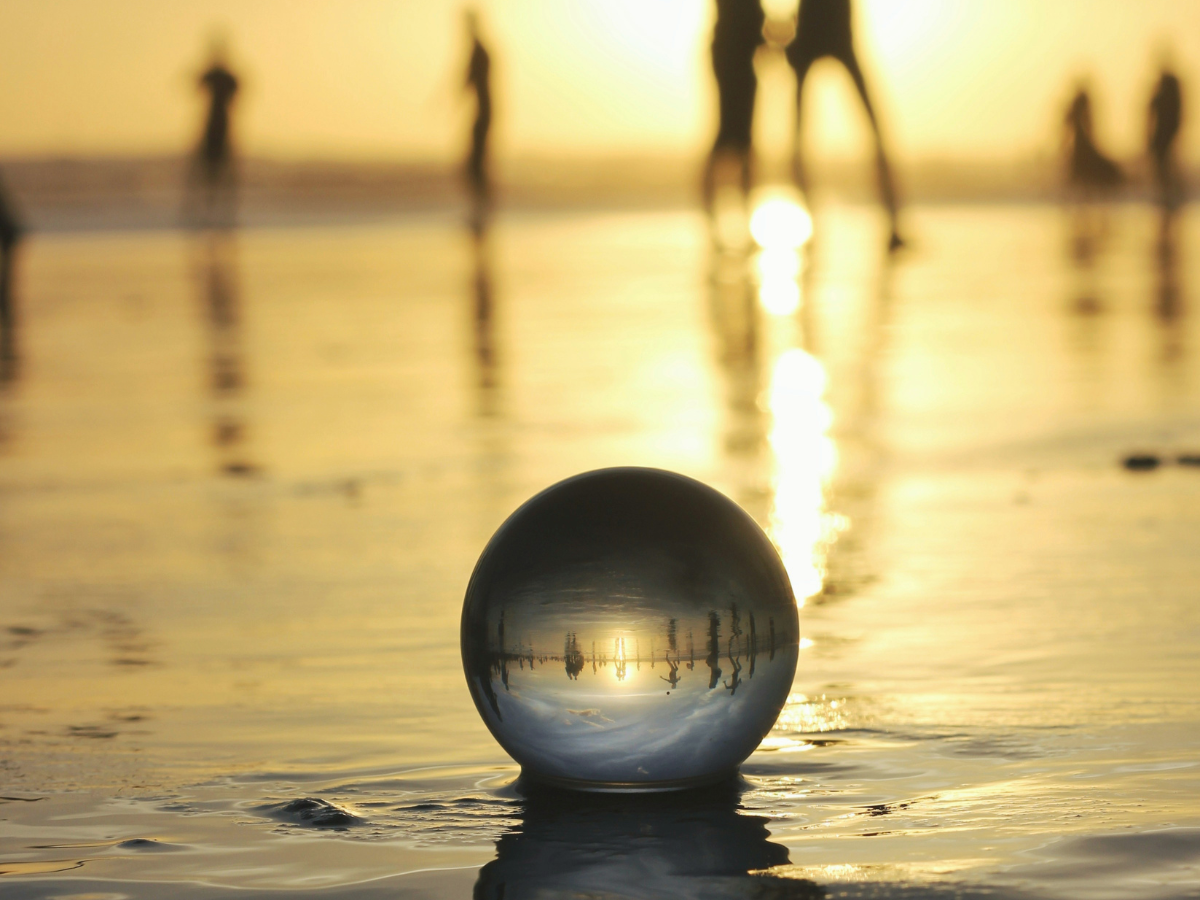 A crystal ball sits in the tide at sunset.  The silhouettes of figures walking by are reflected within its depths.