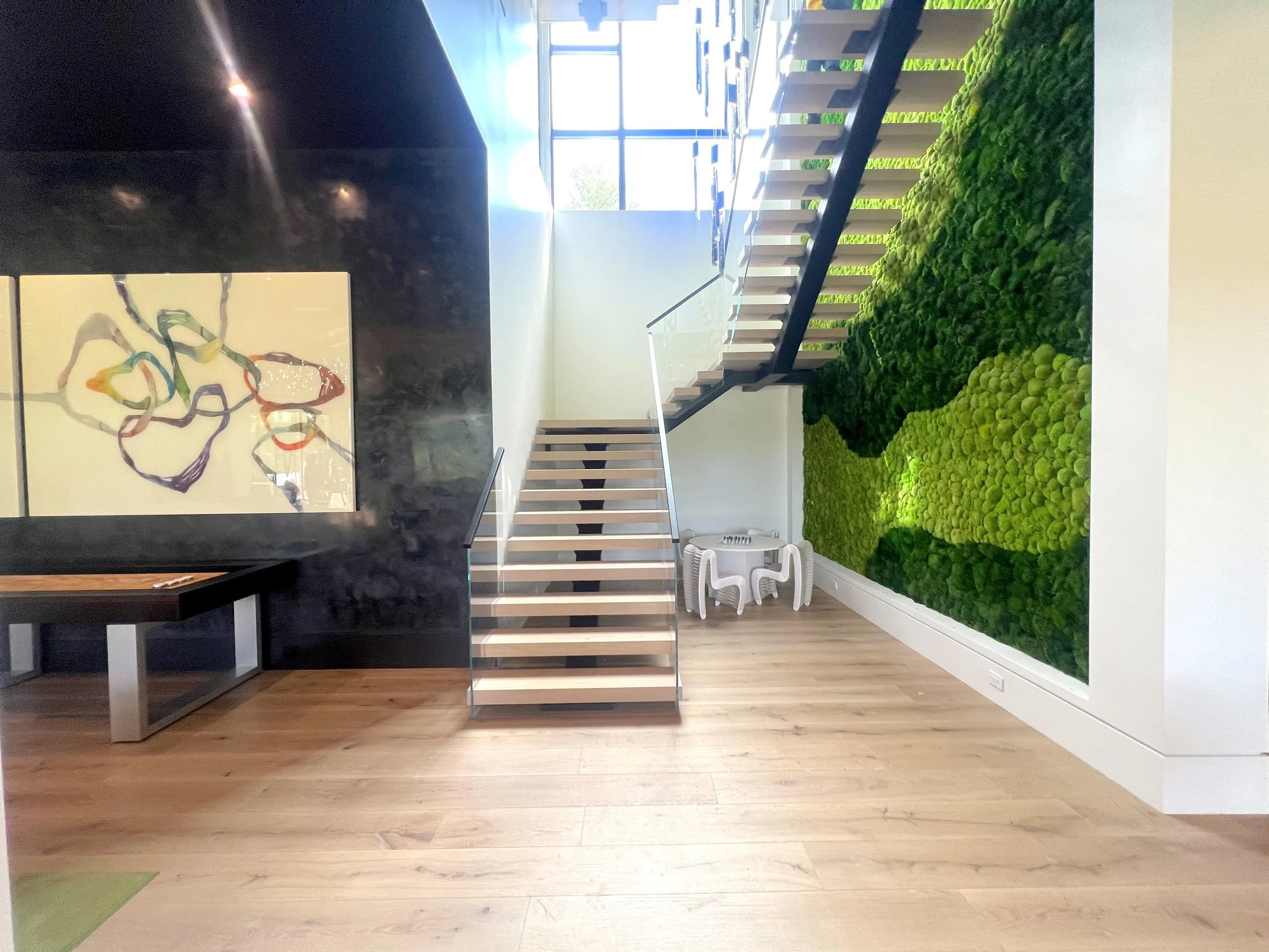 Interior of a modern building with wooden flooring, a black wall with abstract artwork, a staircase with wooden steps and metal railings, a green moss wall, and a small white table with chair-shaped stools.