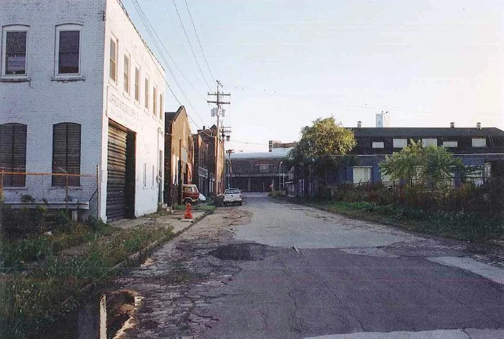 A quiet urban street scene with worn pavement, a white building with closed shutters on the left, a car parked further down, utility poles and wires overhead, a tree on the right side, and a cloudy sky.