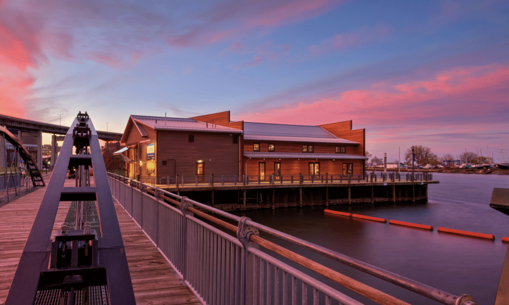 A waterfront scene at sunset with a wooden building on a pier, a curved boardwalk, and sailboats in the background under a colorful sky.