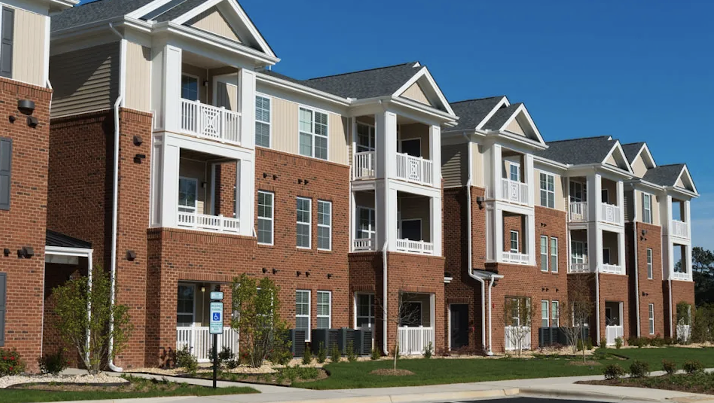 A modern multi-story apartment building with red brick and beige siding, featuring white balconies, windows, and gabled roofs, with landscaped grass, trees, and a sidewalk in the foreground under a clear blue sky.