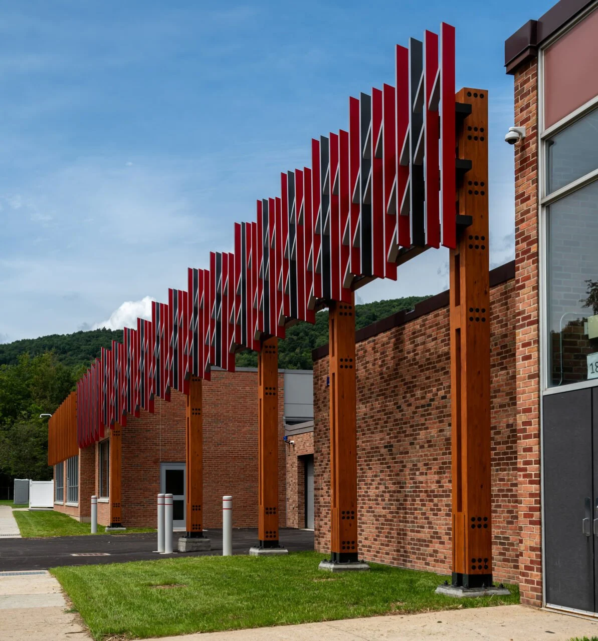 Modern building with red and black vertical metal panels on wooden supports along a brick wall and sidewalk.