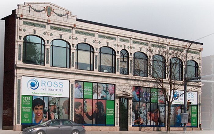 Two-story building with large arched windows, rainbow decorations, and a sign for Ross Eye Institute on the front. Several posters and advertisements are displayed in the windows, and a car is parked in front. A leafless tree is in the sidewalk.