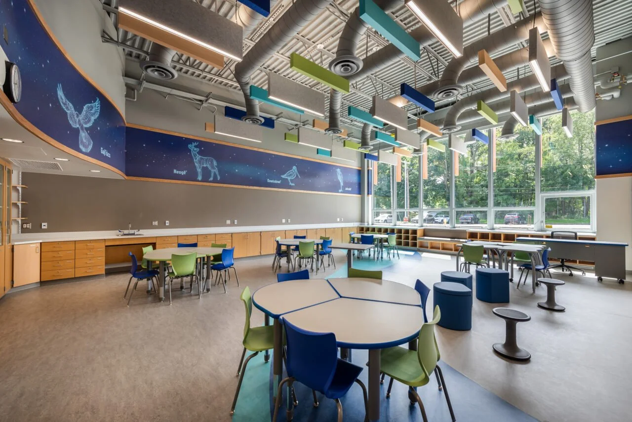 Empty classroom with colorful tables and chairs, large windows, and planets and animals illustrations on a blue space-themed mural.