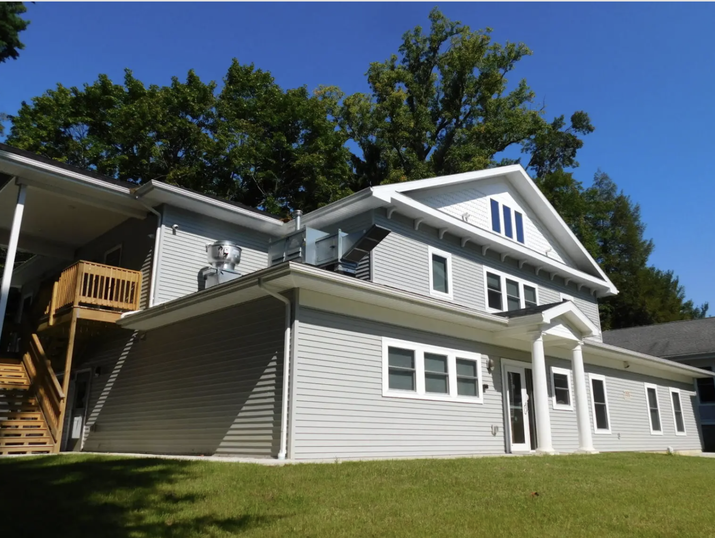 Back view of a gray, multi-story house with white trim, a small balcony with a wooden railing, and a lawn with green grass, under a clear blue sky with trees in the background.