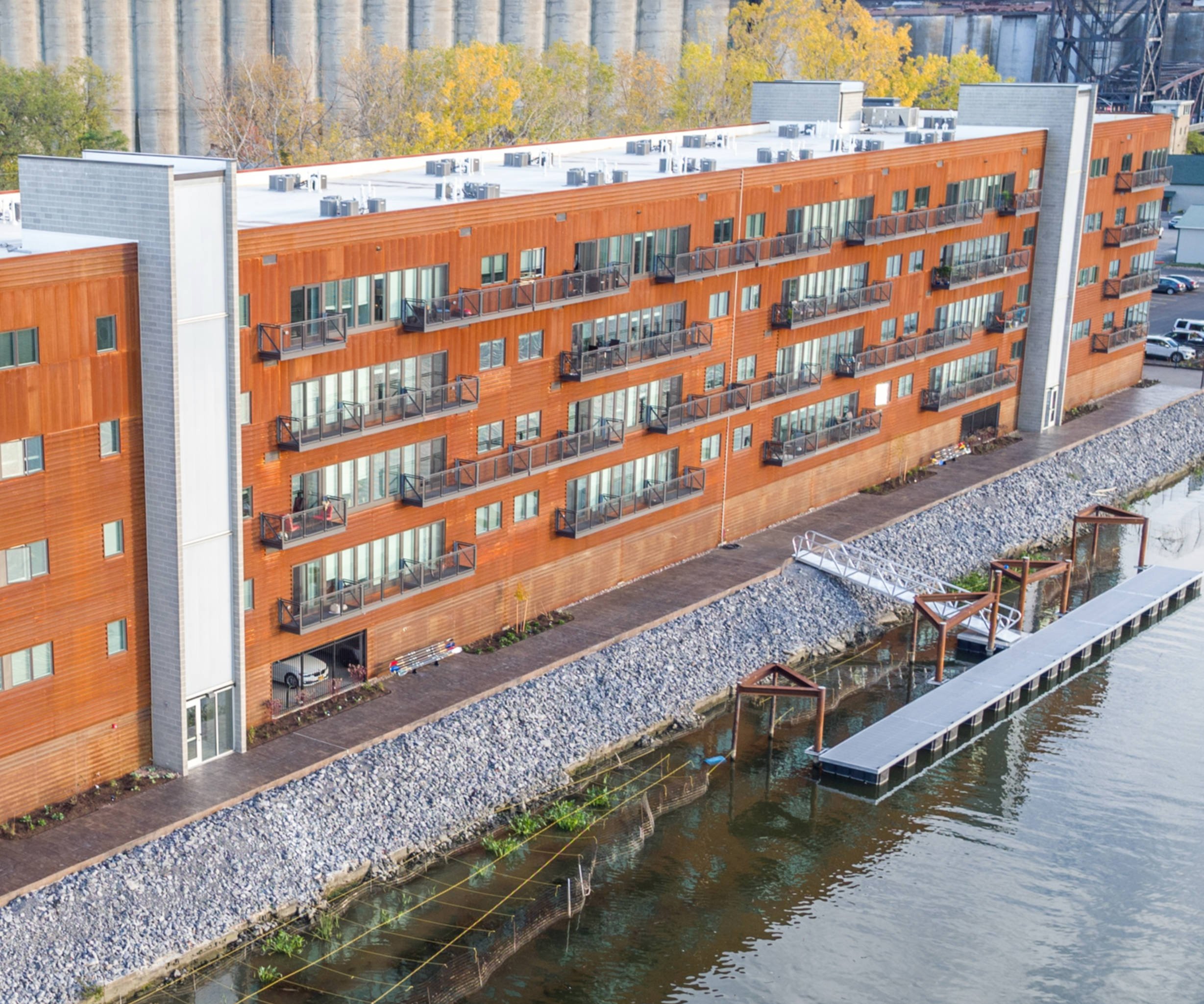 A multi-story residential building with wooden exterior siding, balconies, and a flat roof, situated near a waterway with a rocky shoreline and a walking path.