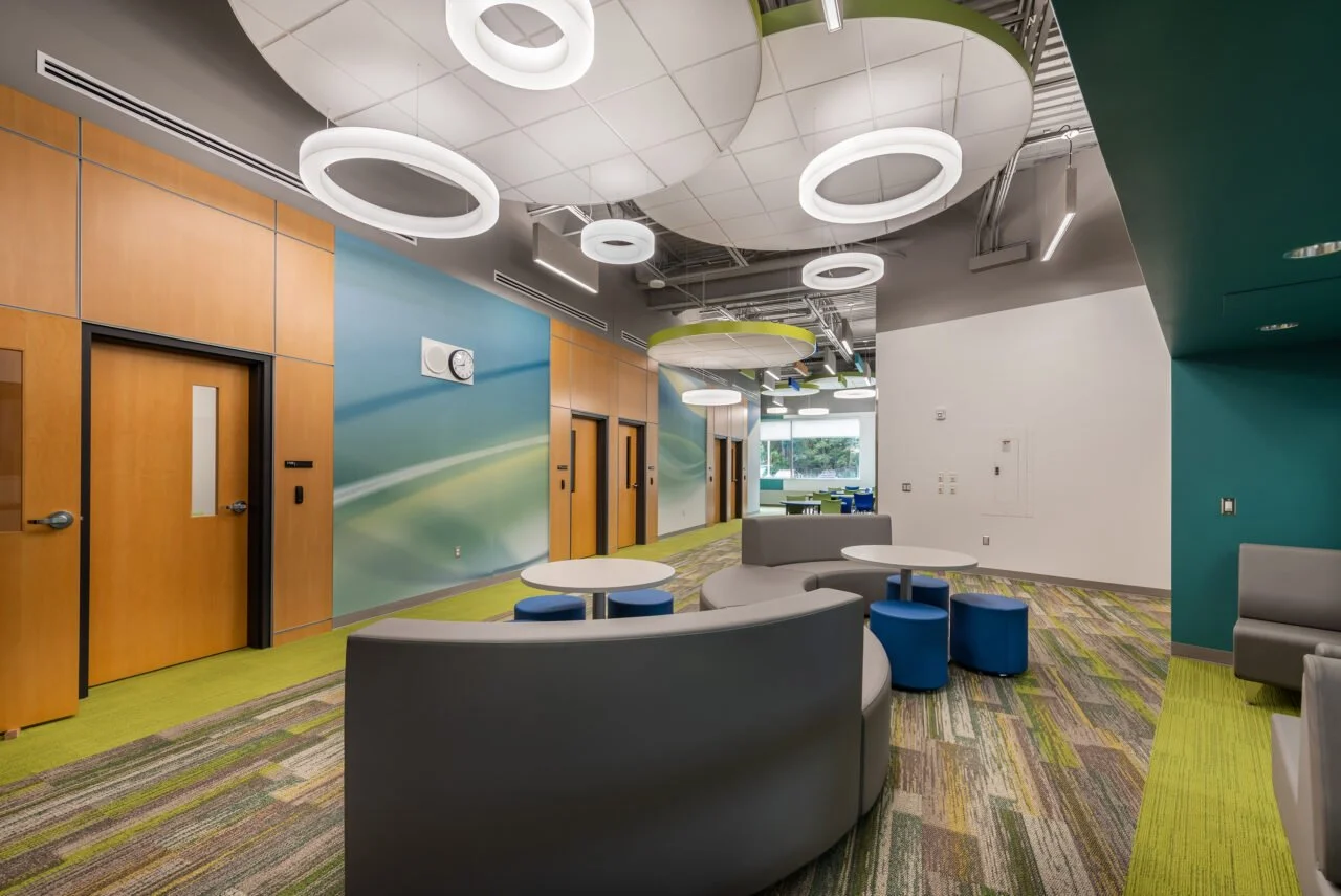 Modern office lounge area with colorful walls, circular lighting fixtures, green, blue, and gray furniture, and multicolored carpet.