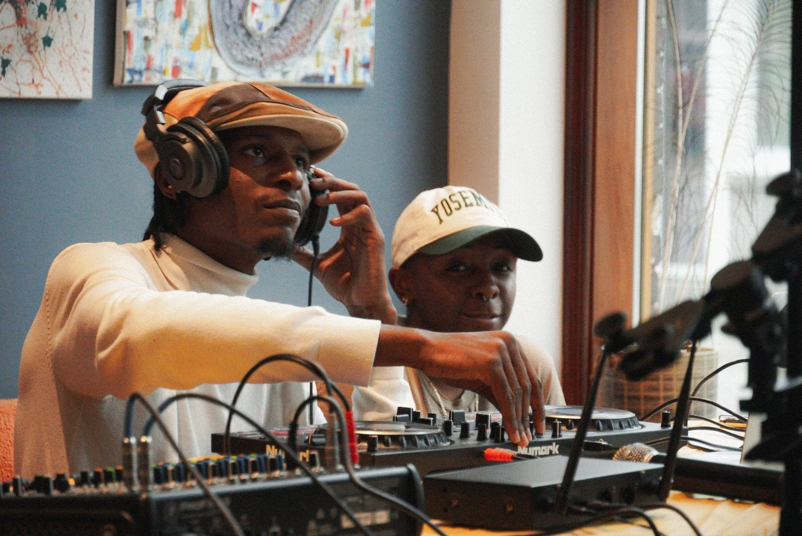 Two people, a man and a woman, are operating DJ equipment. The man is wearing a hat and headphones, and the woman is wearing a cap. They are inside a room with artwork on the wall and a window with natural light.