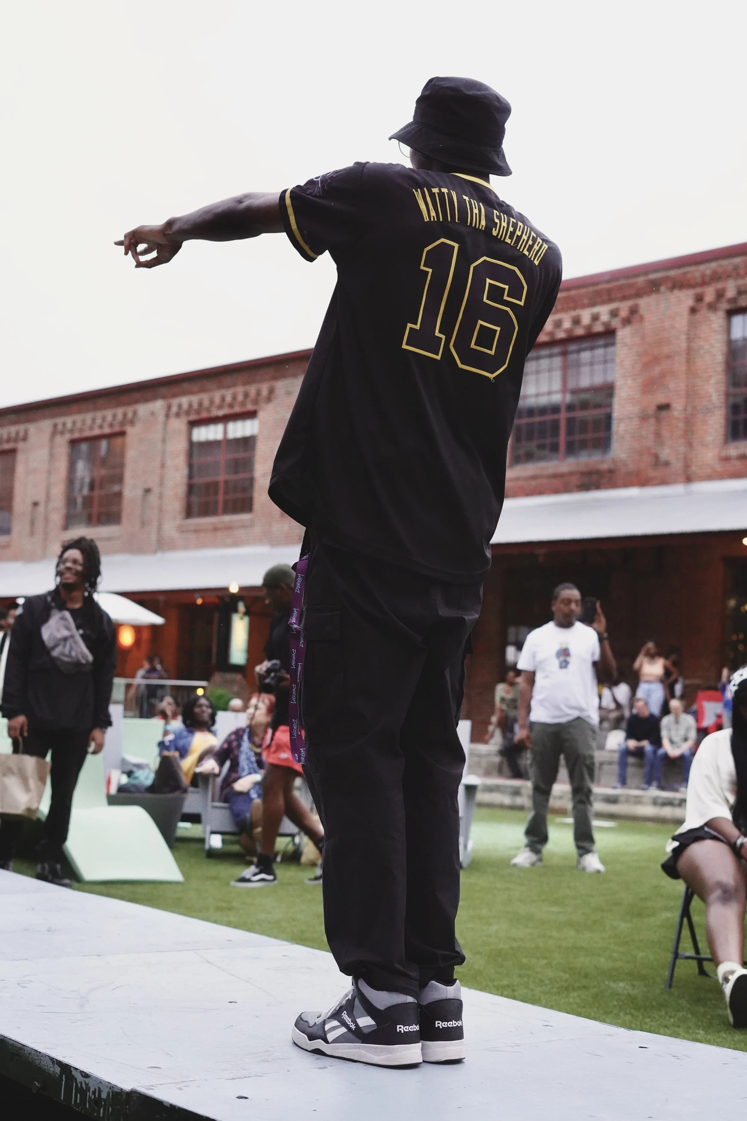 A man wearing a black bucket hat and black sportswear with the words 'Waiting for Shepherd' and the number 16 on the back, standing outdoors on a platform, with people sitting and standing around in an urban courtyard setting.