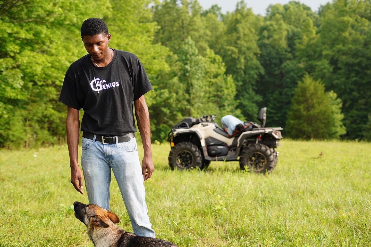 A young man wearing a black T-shirt and light blue jeans standing outdoors on a grassy field, looking down at a brown and black dog sitting beside him; in the background, there is an ATV and a person bent over near it with green trees and foliage surrounding them.