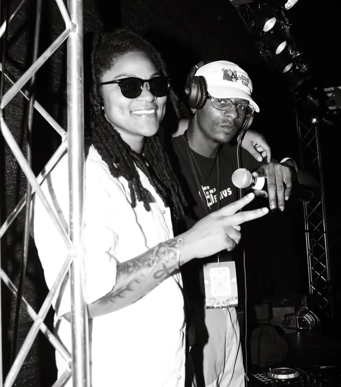 A woman with dreadlocks, wearing sunglasses and a white shirt, smiling and making a peace sign with her hand; a man with glasses, headphones, and a cap, standing next to her, holding a microphone and pointing at the camera; both at a DJ booth with audio equipment in a dark setting.