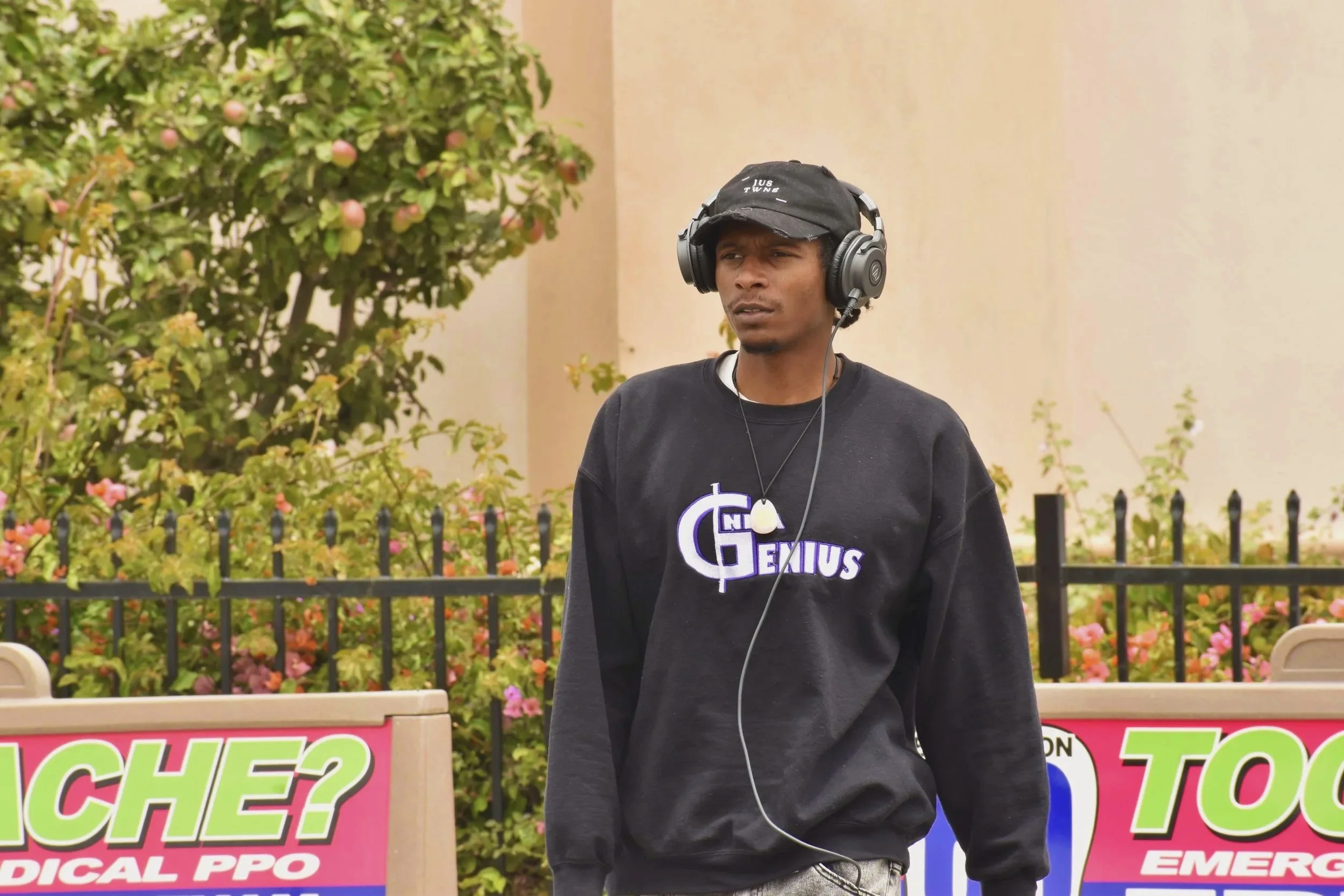 A man wearing a black hoodie with 'Genius' written on it, black cap, and headphones, standing outdoors near a colorful sign and bushes.