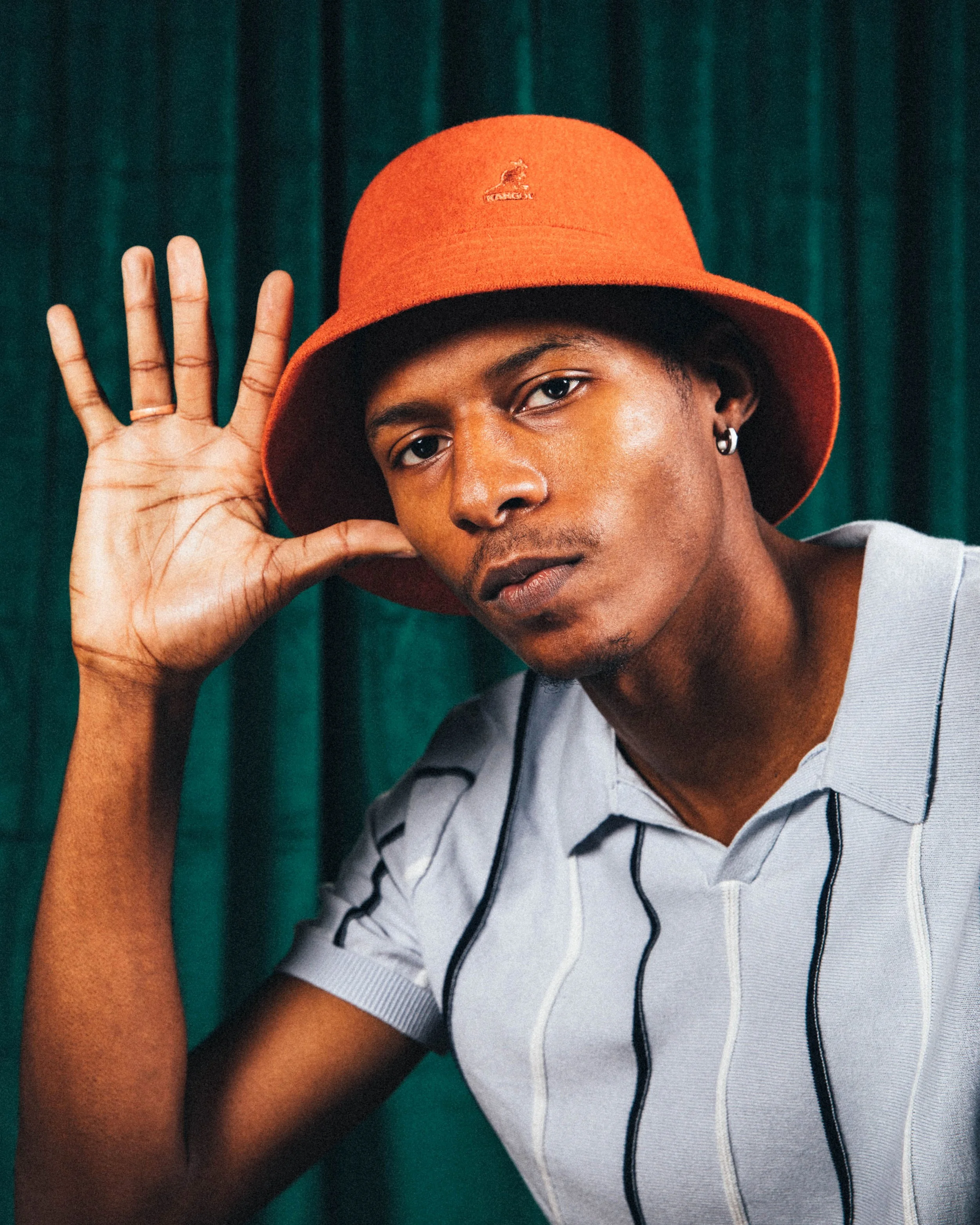 A young man with dark skin, wearing a red bucket hat and a light gray, striped shirt, posing against a dark green background. He is touching his face with his right hand and looking directly at the camera.