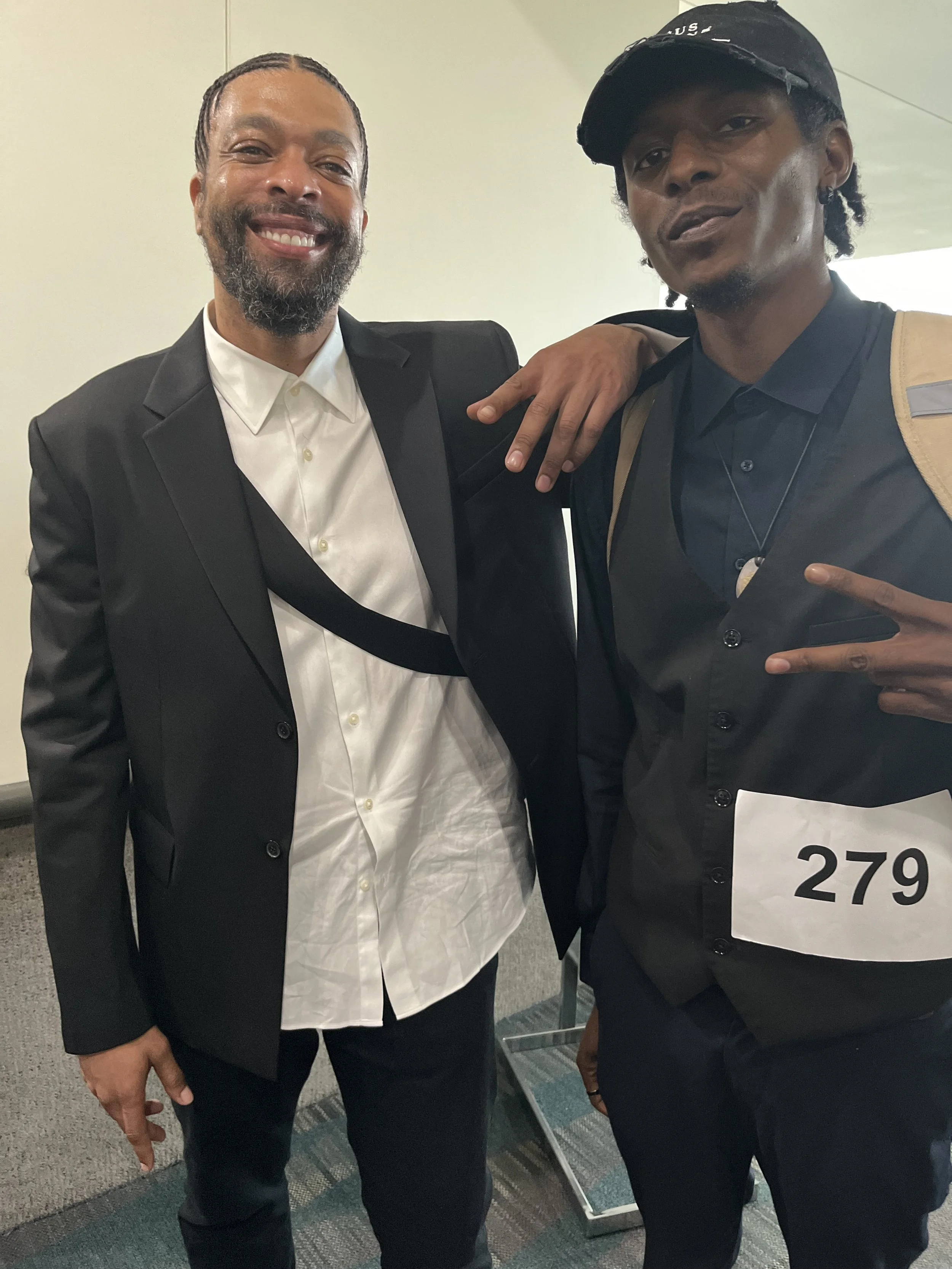 Two men posing together, one is smiling with his arm over the other's shoulder, both dressed in black suits. The man on the right is wearing a cap and making a peace sign, with a numbered badge reading '279' on his chest.
