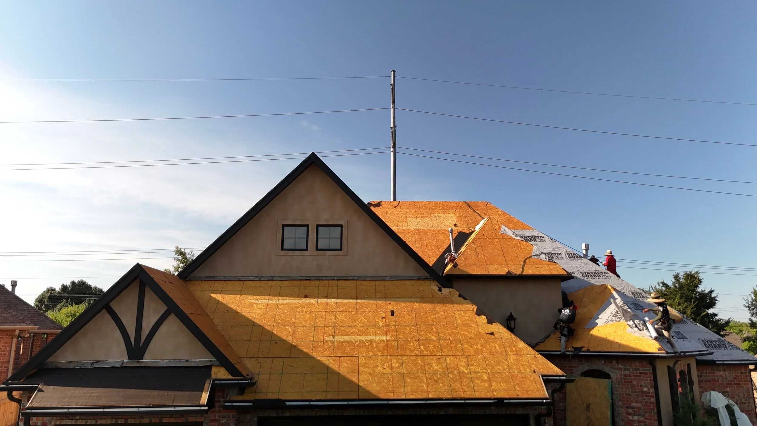 Construction workers installing roofing materials on a house with orange sheathing and underlayment, during daytime with clear sky.