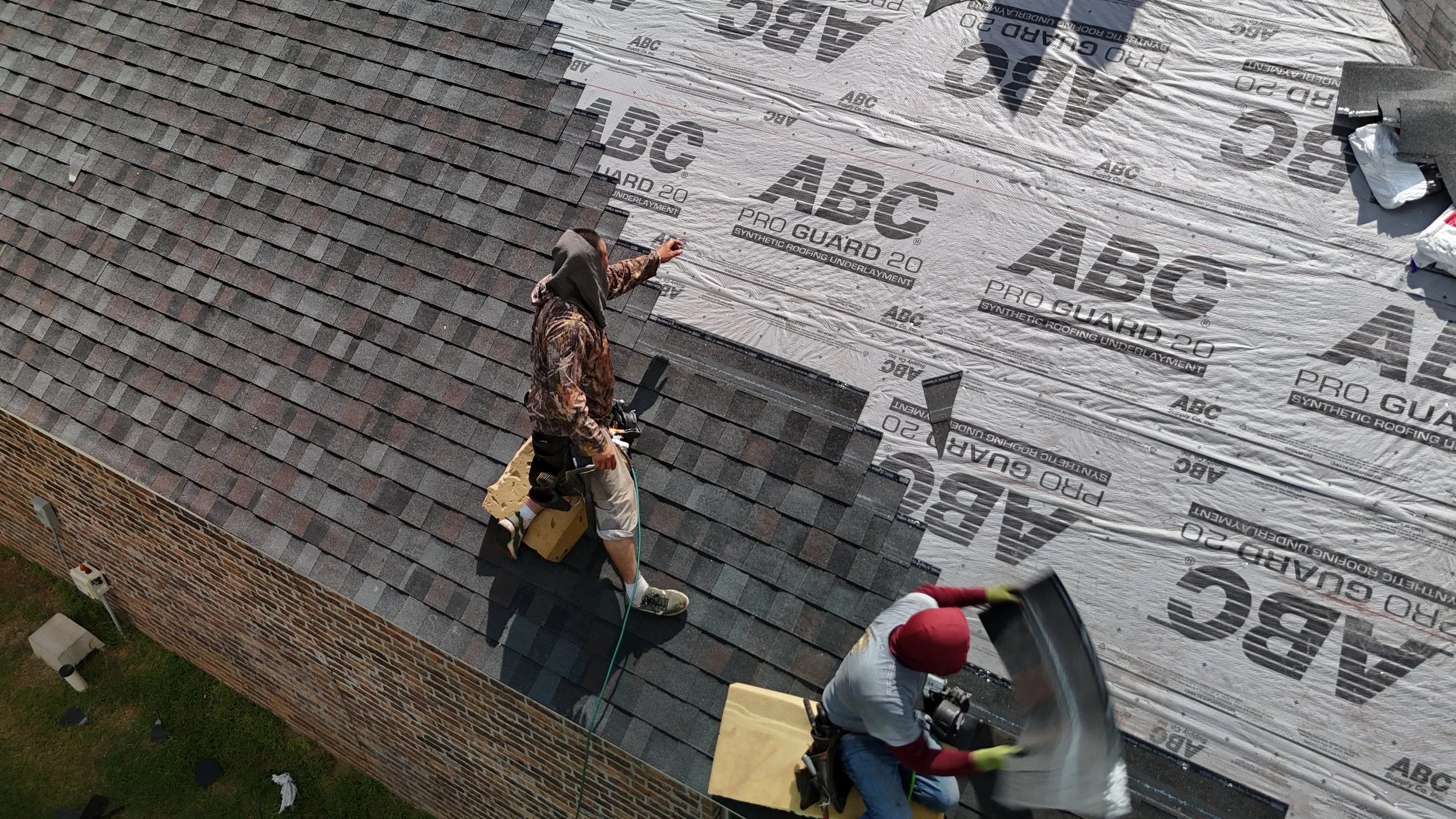 Two workers installing asphalt shingles on a roof. One worker is using a blowtorch, while the other is handling a bundle of shingles.