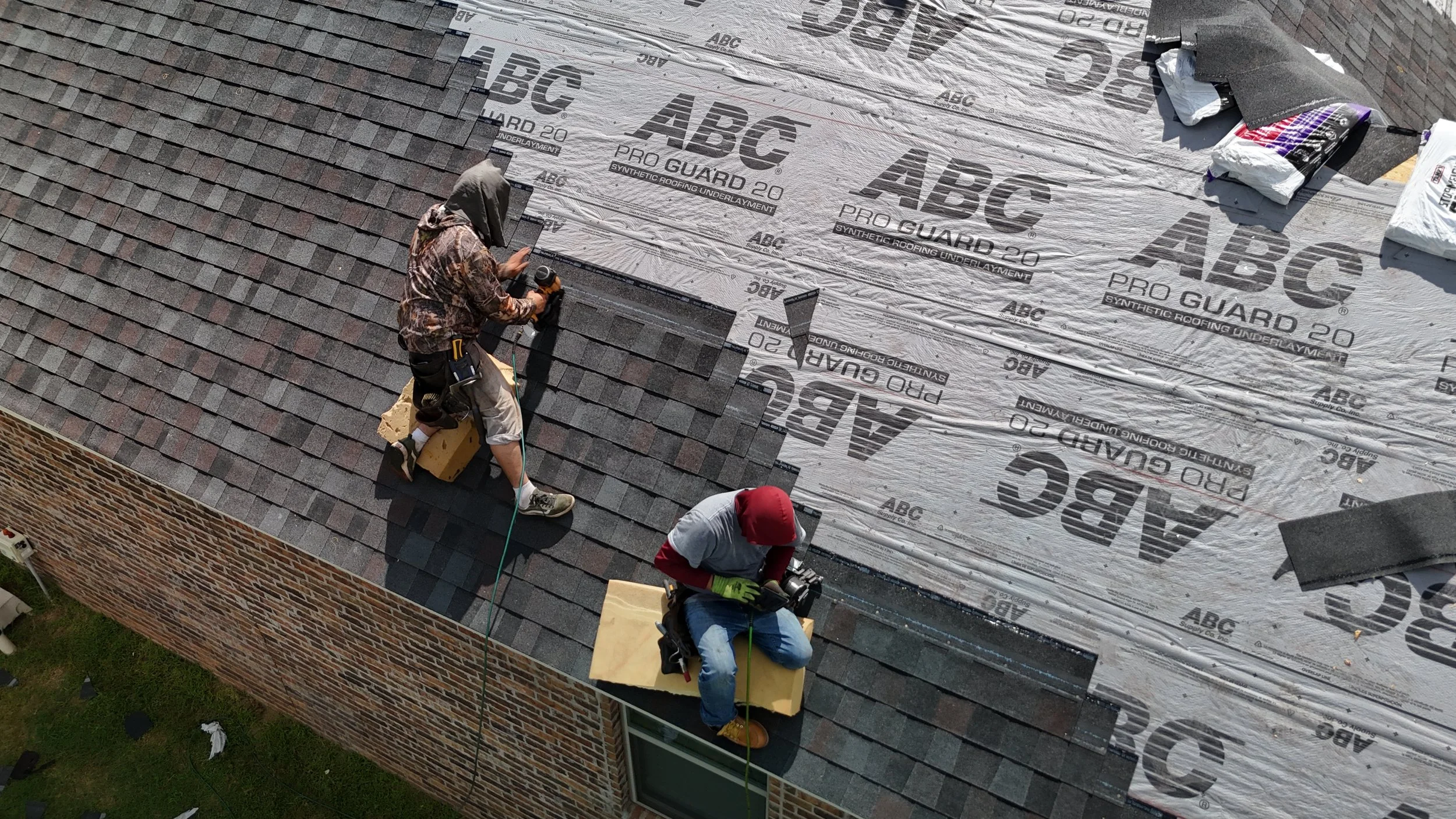 Two workers installing shingles on a roof, with parts of the roof covered in synthetic roofing underlayment labeled 'ABC PRO GUARD 20'. One worker is kneeling, and the other is sitting, both wearing safety gear and using tools.