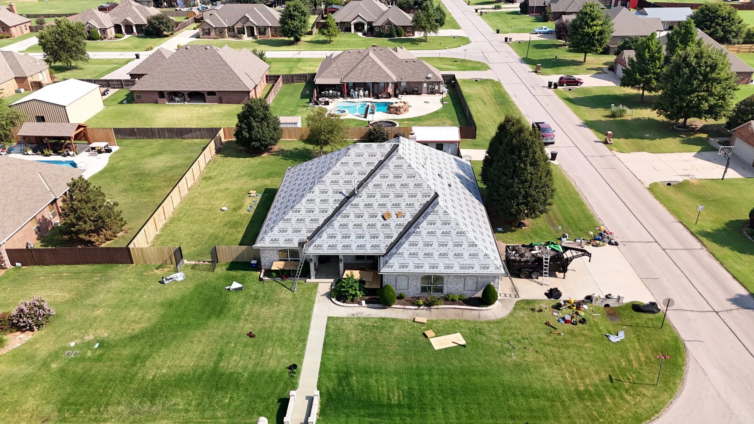 A house under construction with a roof being built, surrounded by a landscaped yard with trees, lawns, and nearby houses in a suburban neighborhood.