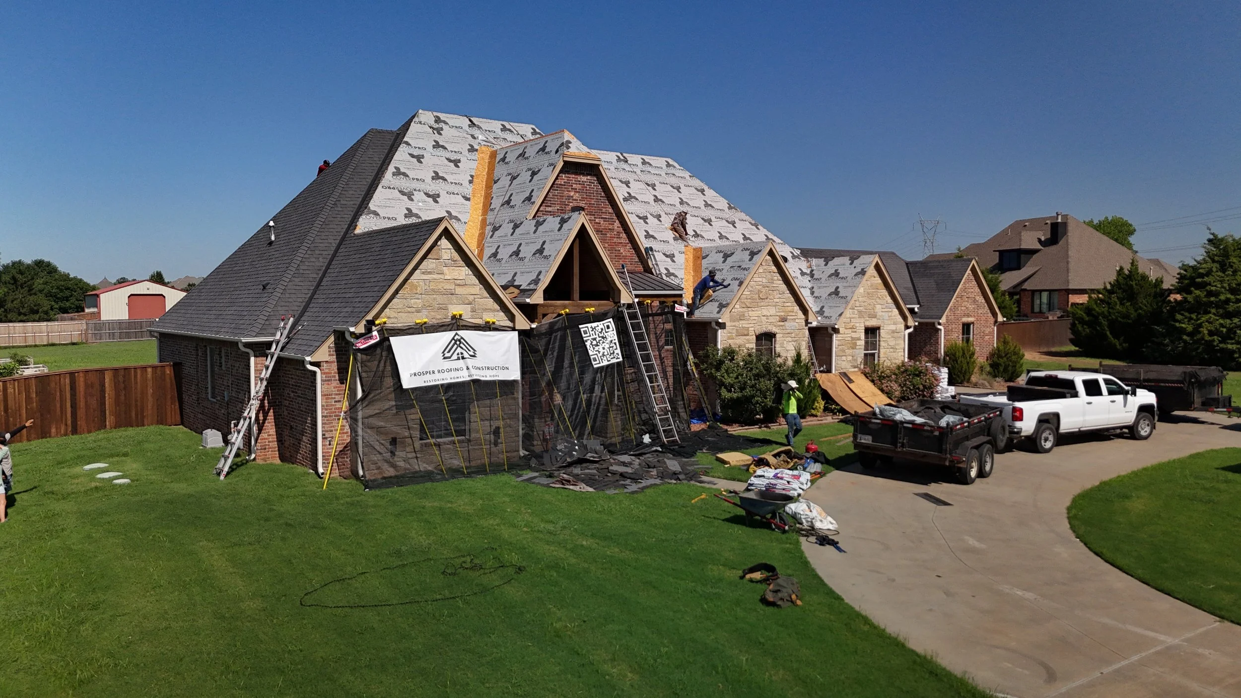 House under construction with workers on the roof, roofing materials, and trucks parked in the driveway.