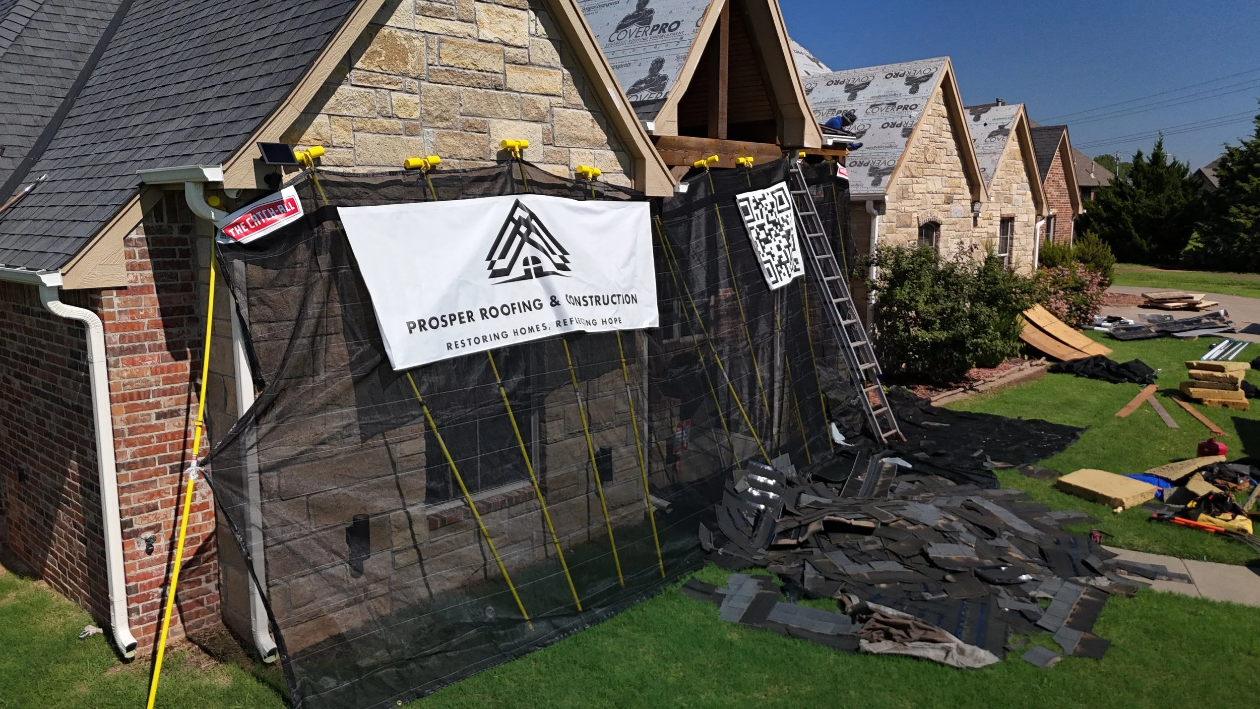 Construction site of a house showing partly built walls with roofing work in progress, ladders, construction materials, and equipment on a grassy yard under a clear blue sky.