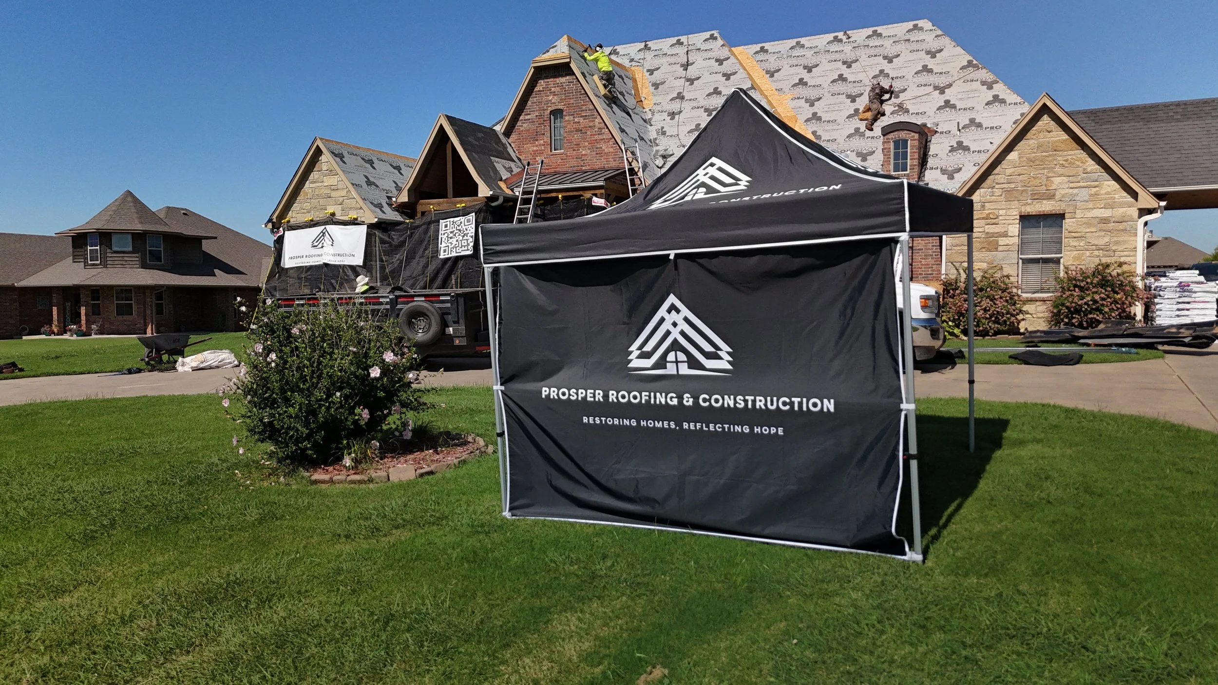 Construction workers on the roof of a house under roofing renovation, with a black tent in front displaying 'Prosper Roofing & Construction' logo and slogan.