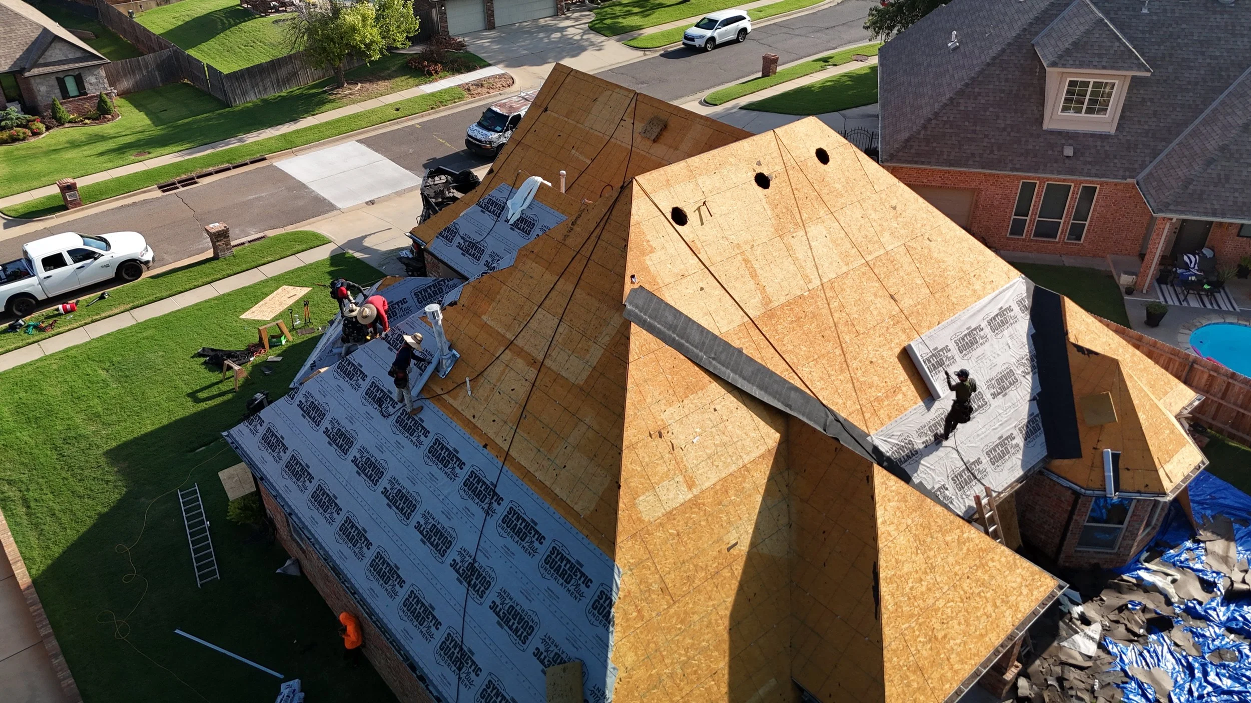 Aerial view of a house under construction with workers on the roof installing roofing materials. The house is in a suburban neighborhood with neighboring houses, cars parked on the street, a lawn, and a pool visible in the backyard.