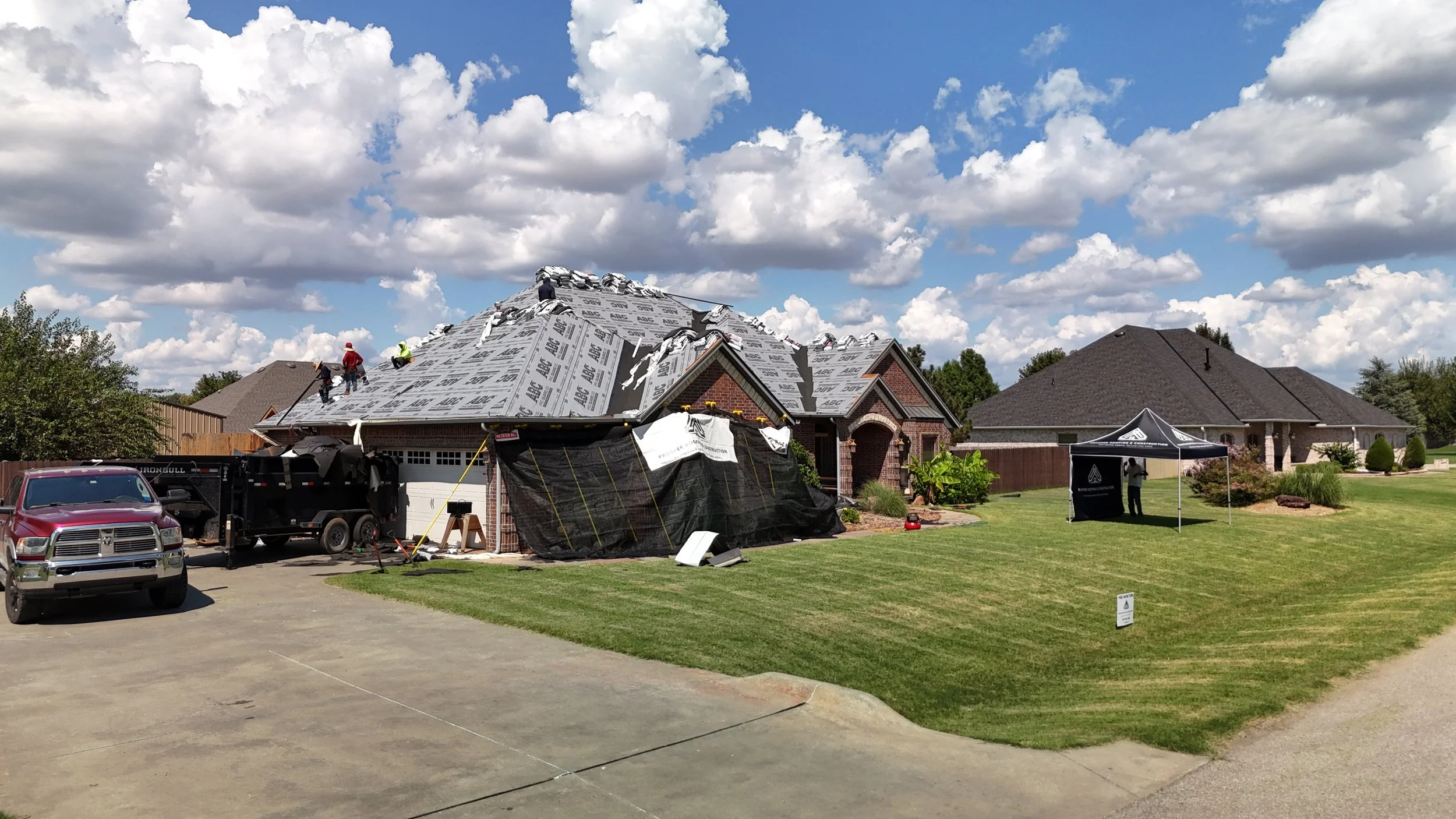 House undergoing roof replacement with workers on the roof, construction equipment, and a temporary tent set up in the yard.