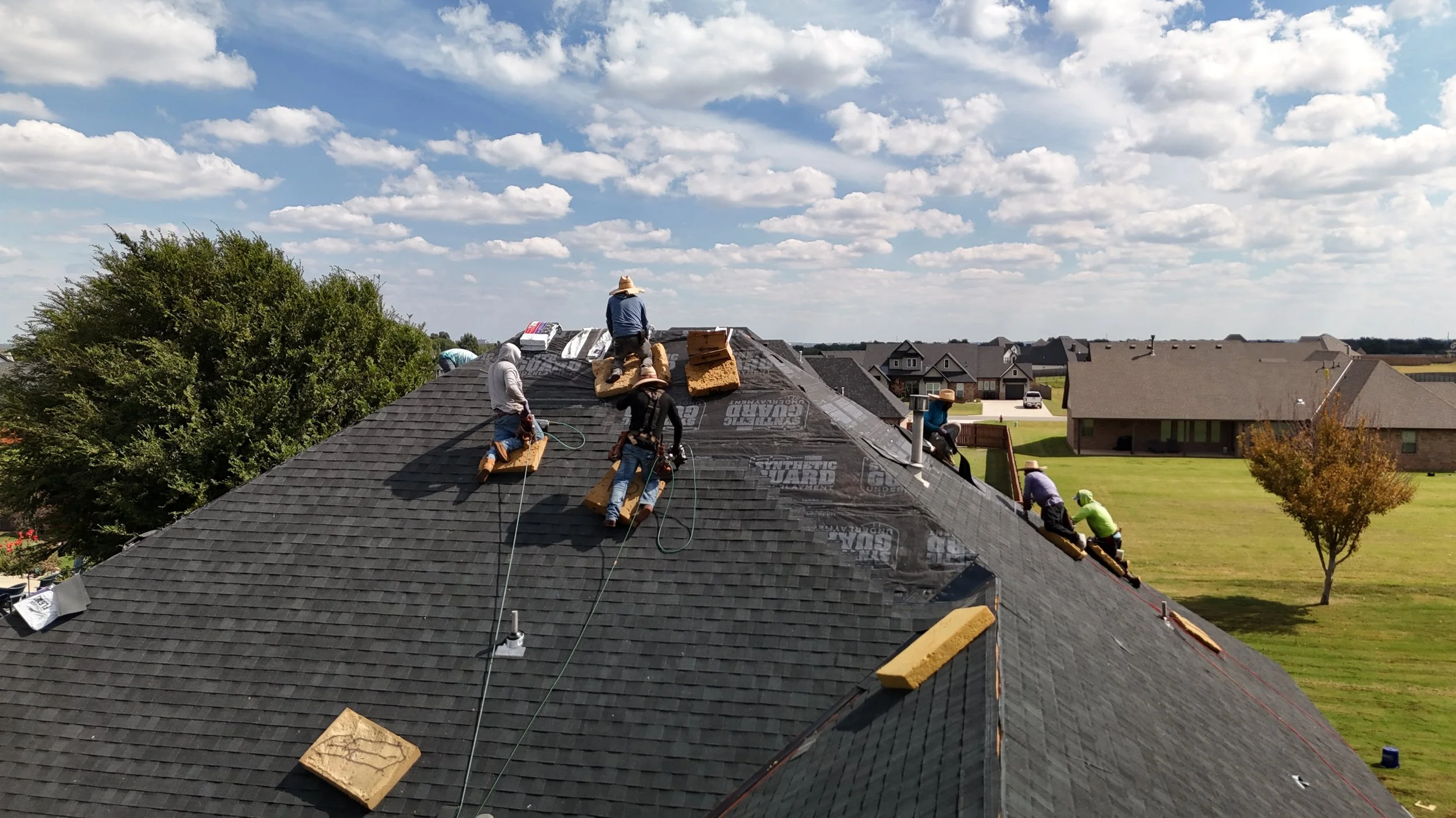 Roofers working on a house roof with shingles, some sitting on the roof, others climbing or working near the chimney, under a partly cloudy sky in a suburban neighborhood.