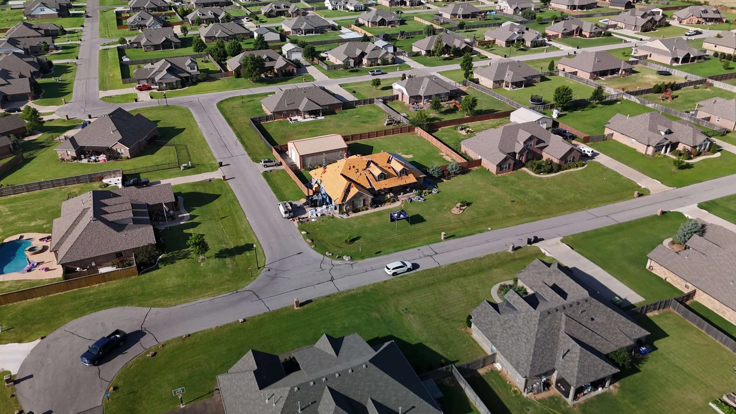 Aerial view of a suburban neighborhood with single-family homes, green lawns, and some swimming pools. One house appears to be under construction with a partially built roof.