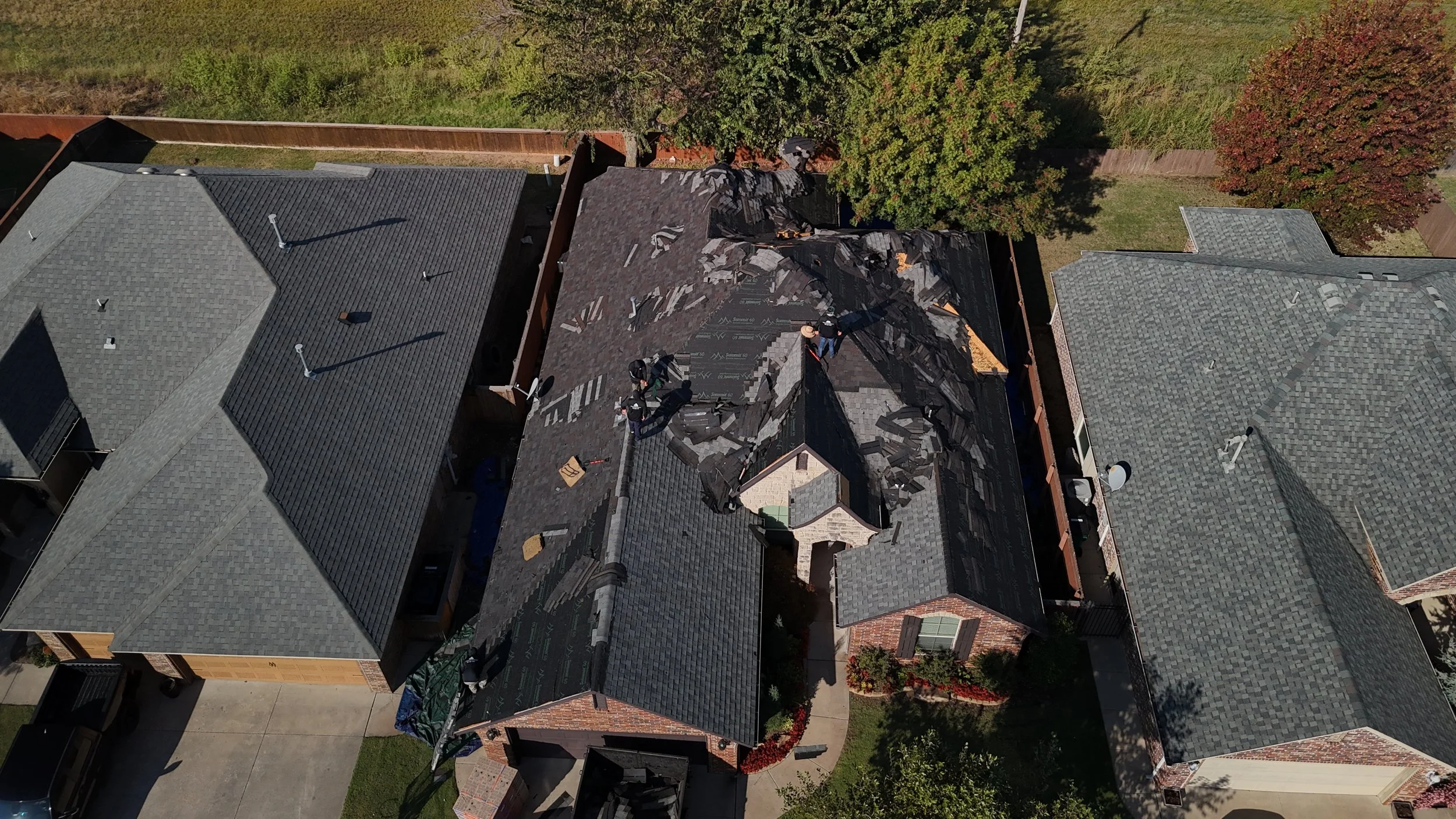 Aerial view of a house with roofing damage as workers repair the roof, surrounded by neighboring houses and trees.