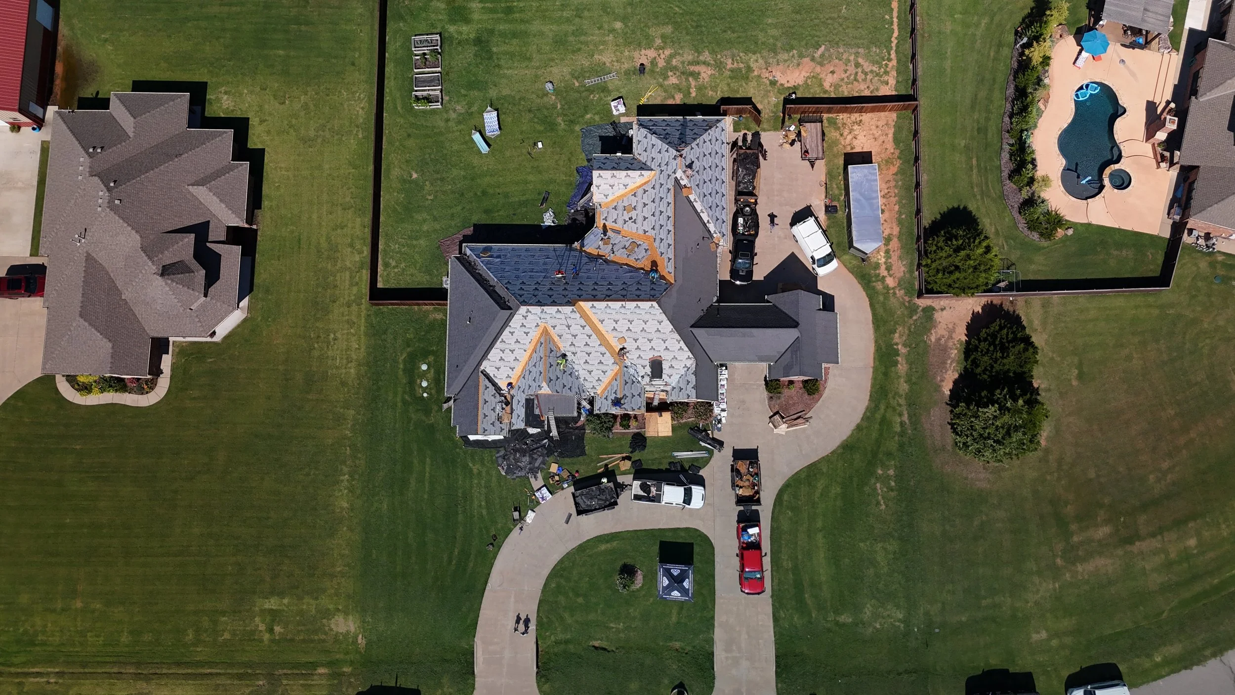 Aerial view of a house under construction showing roofing work, a lawn, parked cars, a backyard with a swimming pool, and neighboring houses.