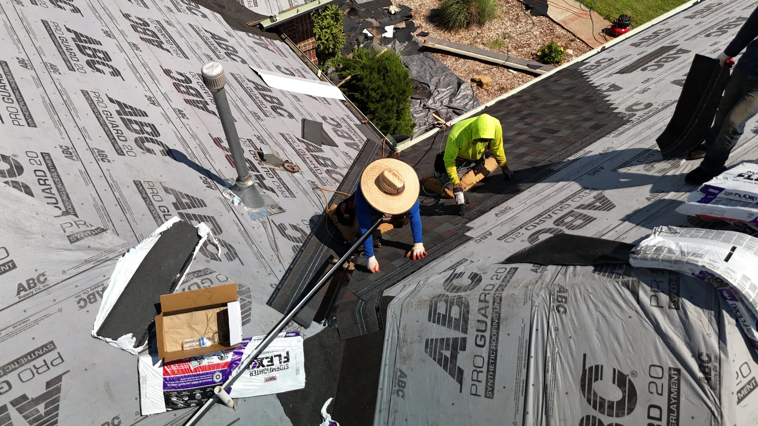 Two workers installing asphalt shingles on a roof, with construction materials and tools around them.