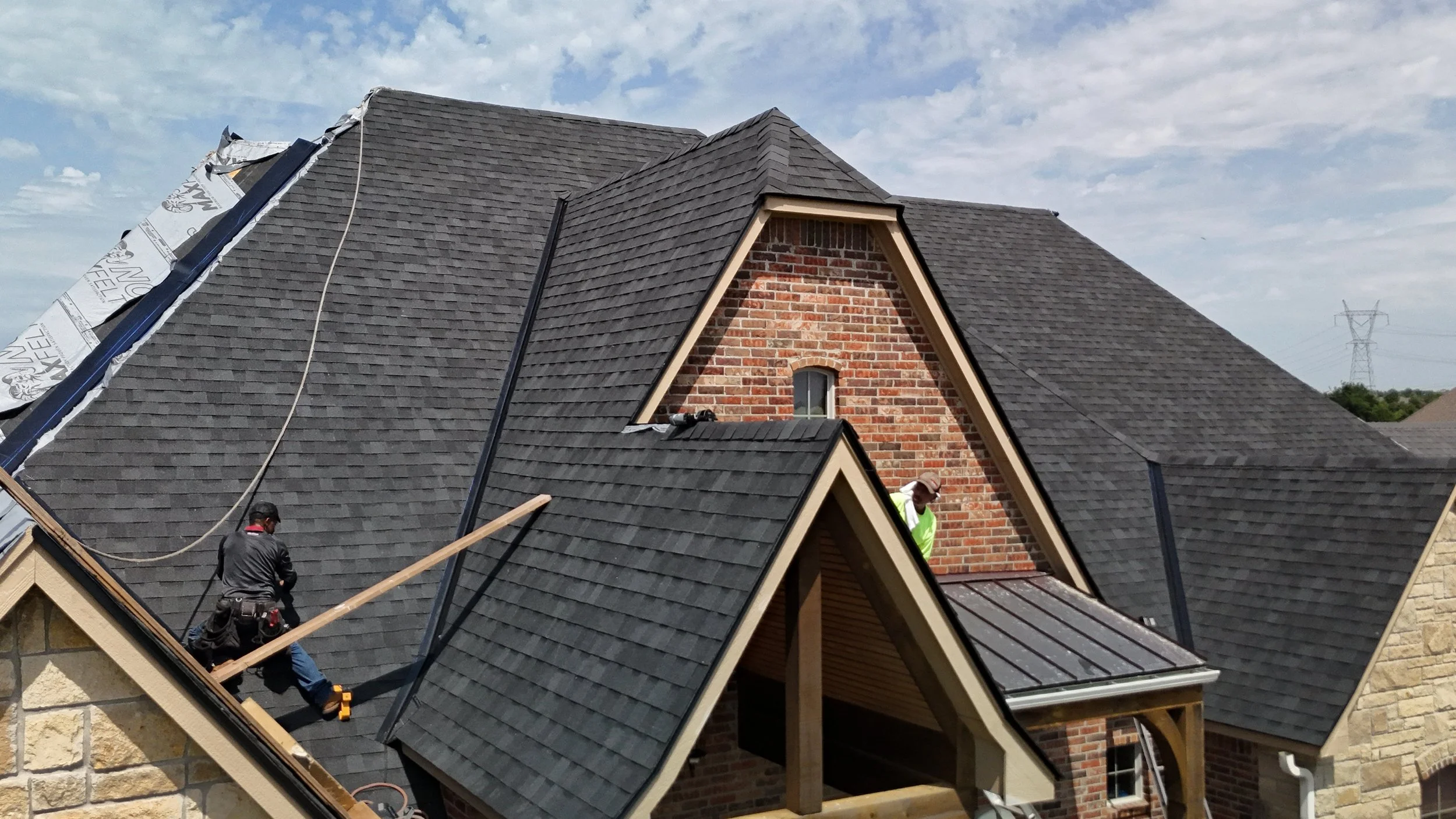 Two workers installing or repairing roofing shingles on a house with a steep roof, partially covered with underlayment, under a partly cloudy sky.