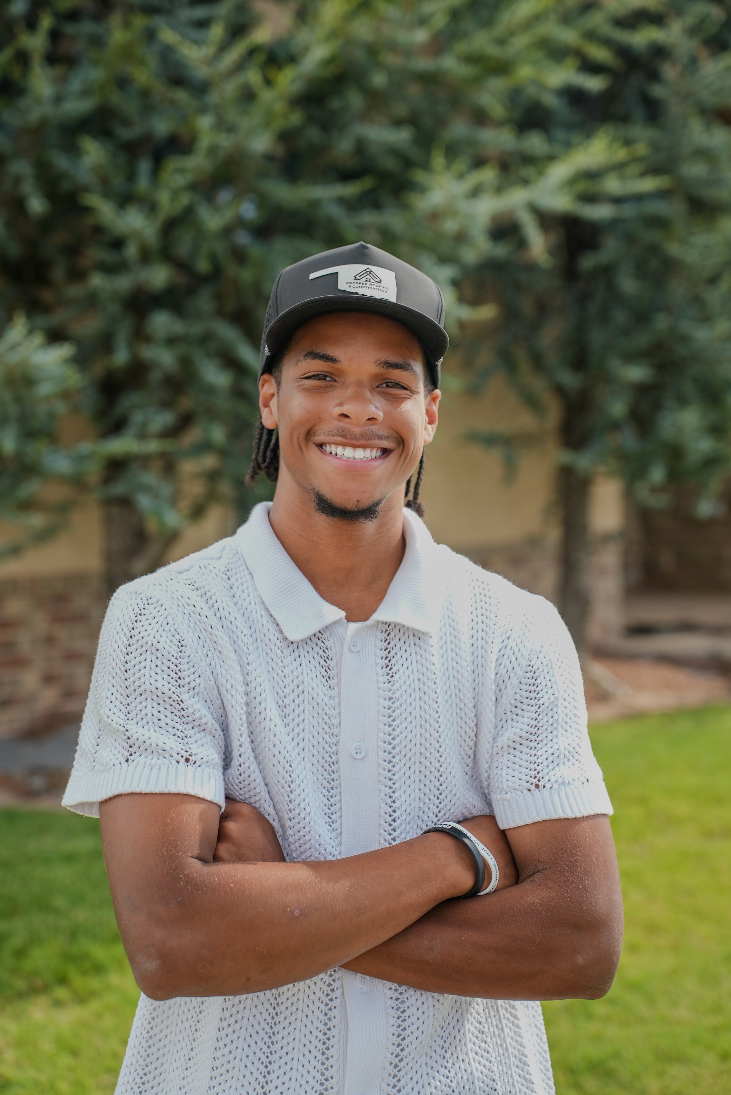 A young man wearing a gray polo shirt and a black baseball cap with a logo, standing outdoors in front of a residential house, smiling with arms crossed.